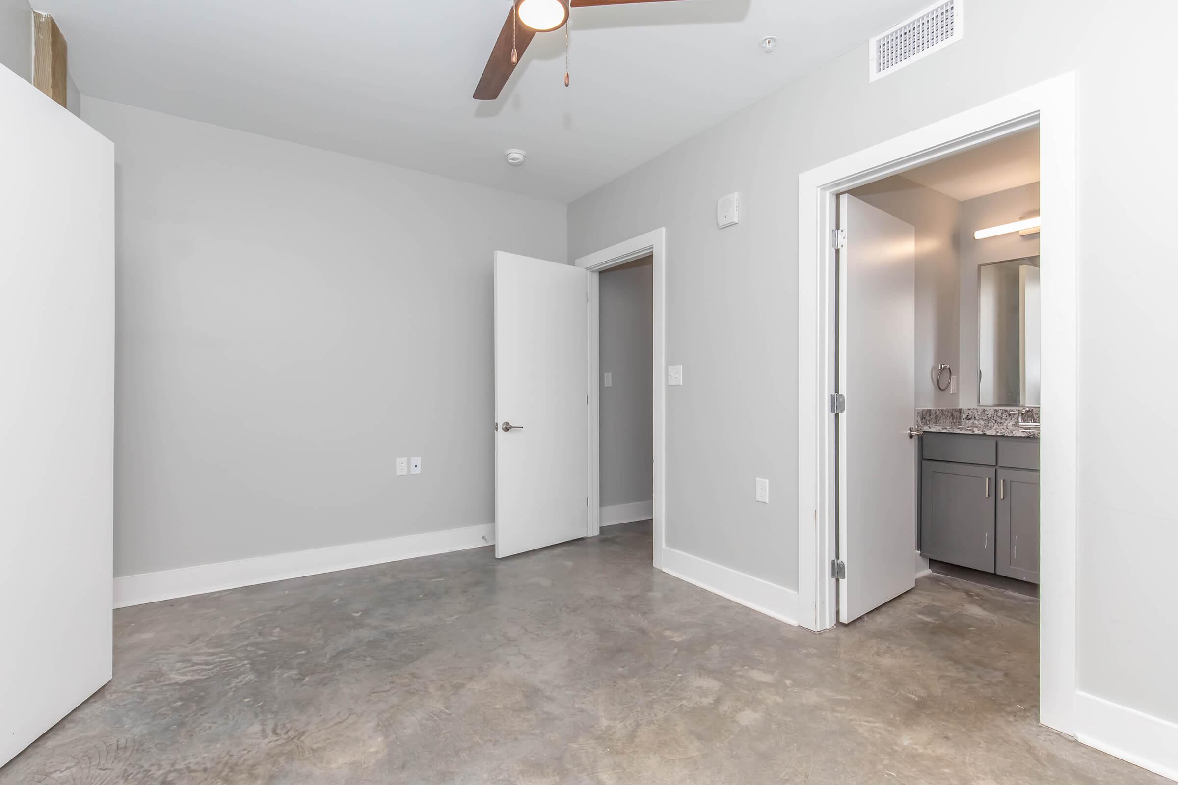 A modern, empty room with gray walls and a ceiling fan. There is a door leading to a bathroom on the right, which features a mirror and a vanity with a granite countertop. The floor is polished concrete, and a closet door is visible on the left side. The space is well-lit with natural light.
