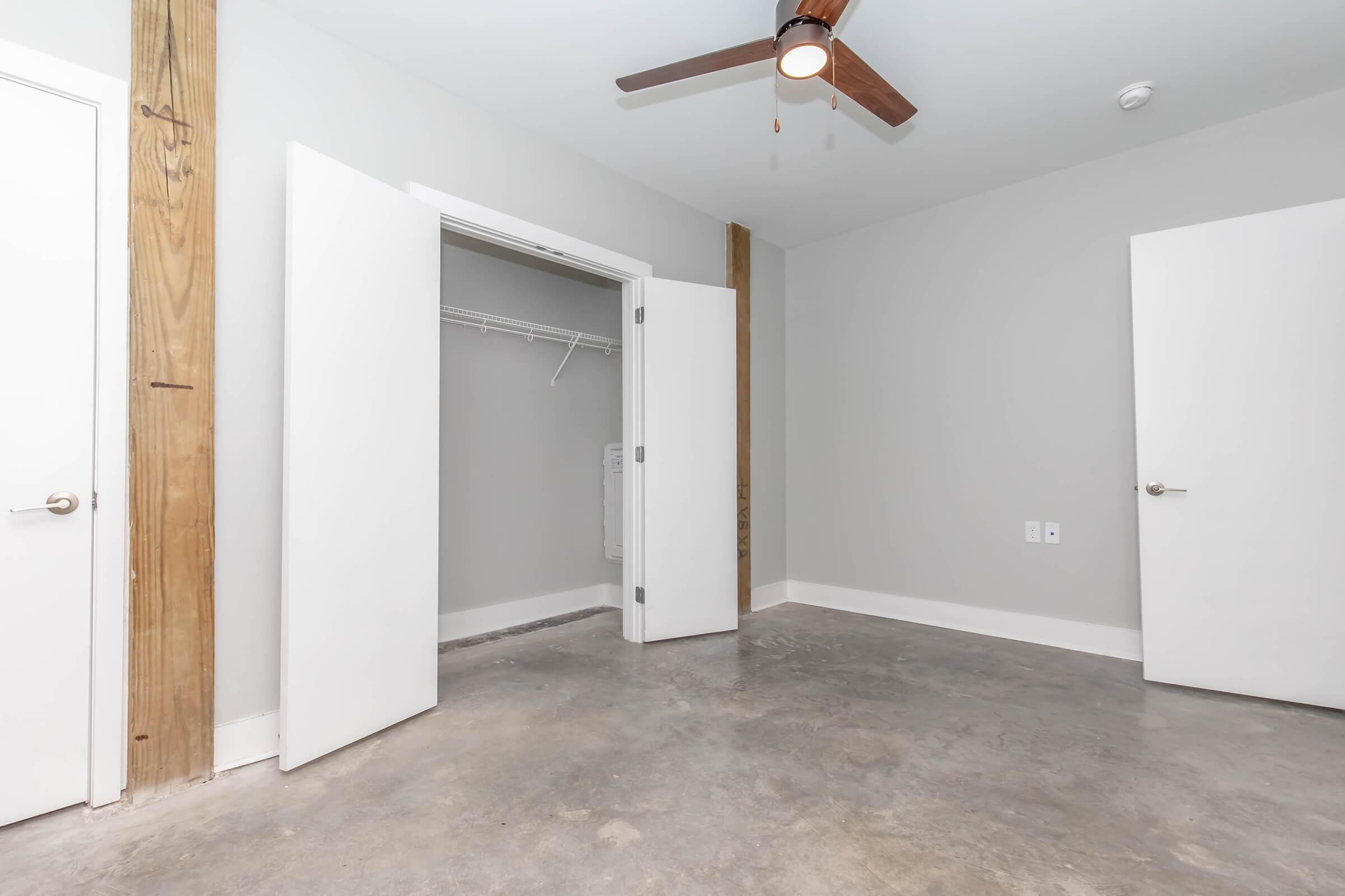 A blank, minimalist room with gray walls and a polished concrete floor. There are two white doors; one leads to a closet with open shelving. A ceiling fan with wooden blades is installed, and the room has a spacious feeling due to the absence of furniture. Natural light brightens the space through the doorway.