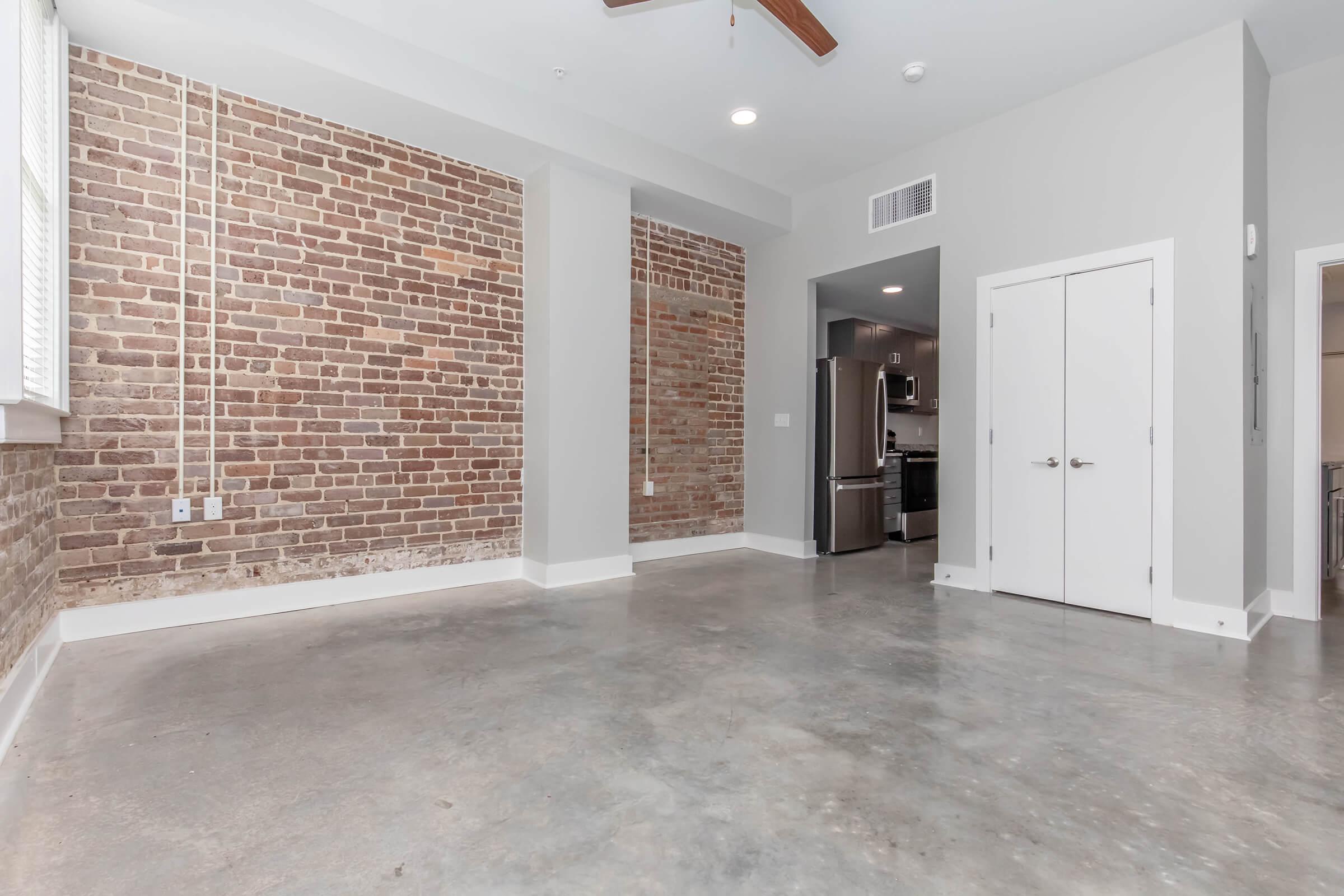 An interior view of a modern apartment featuring exposed brick walls, a concrete floor, and a ceiling fan. In the background, there is a doorway leading to a kitchen with stainless steel appliances. The space is bright with natural light coming from a nearby window.