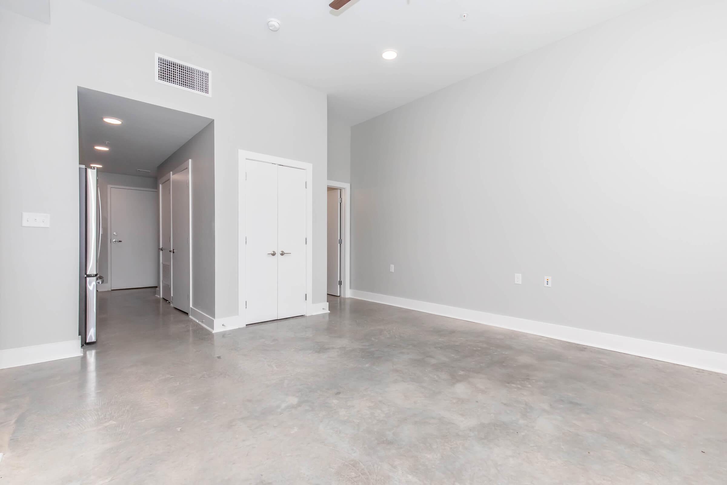 A spacious, empty room with gray walls and polished concrete floors. There are two doorways leading to other rooms, and a sleek stainless steel refrigerator is visible on the left. The room features overhead lights and a ceiling fan, giving it a modern look.