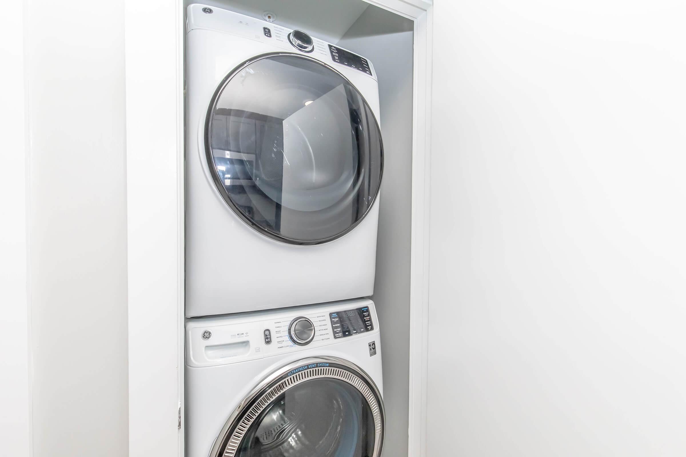 A stacked washer and dryer unit in a small laundry area. The machines are white with a modern design, featuring large circular doors and control panels at the front. The setting is well-lit with white walls, creating a clean and organized appearance.