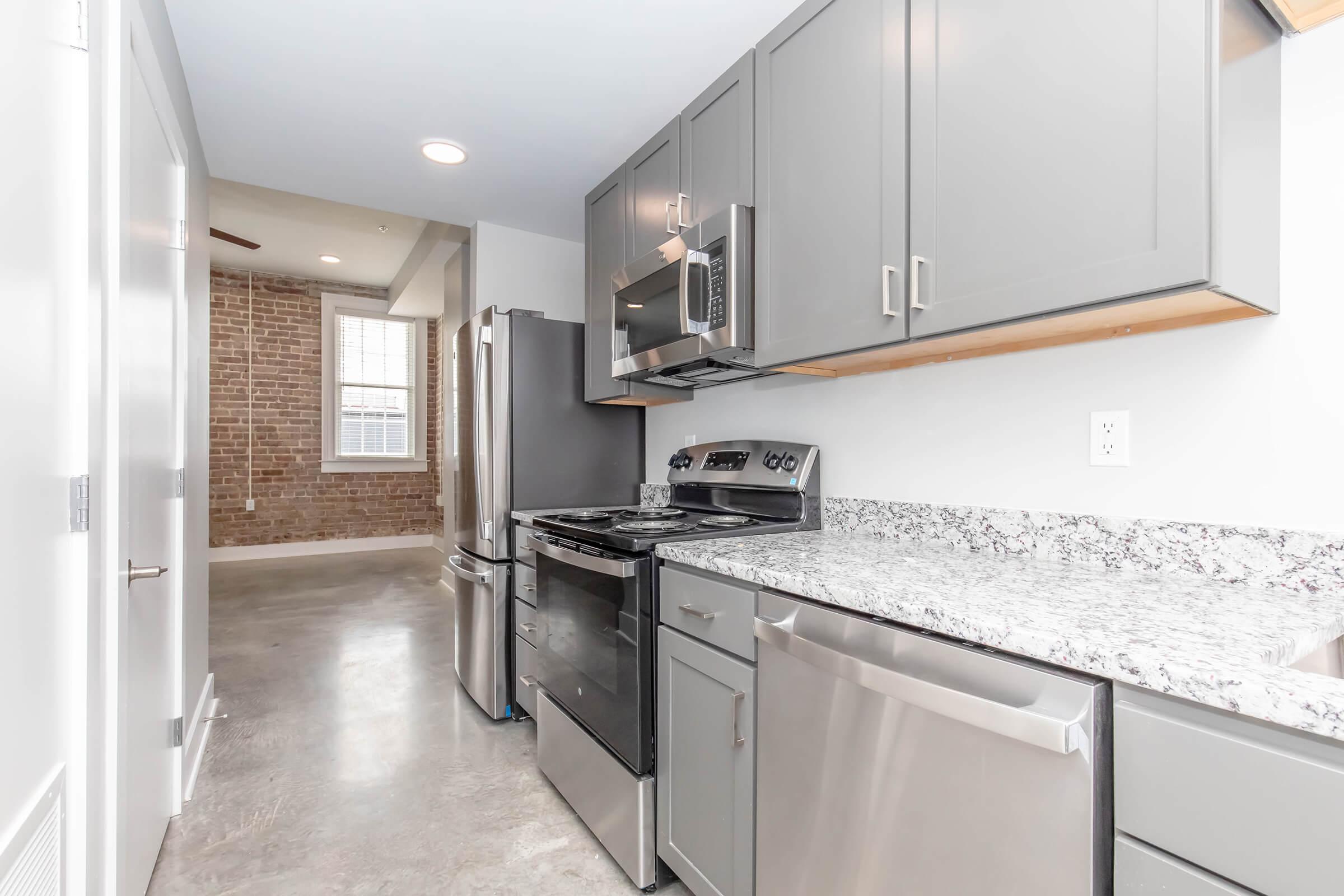 Modern kitchen featuring gray cabinets, stainless steel appliances, and a granite countertop. The space is well-lit with natural light from a nearby window and has exposed brick walls, providing an industrial feel to the setup. The floor is a polished concrete finish, complementing the contemporary design.