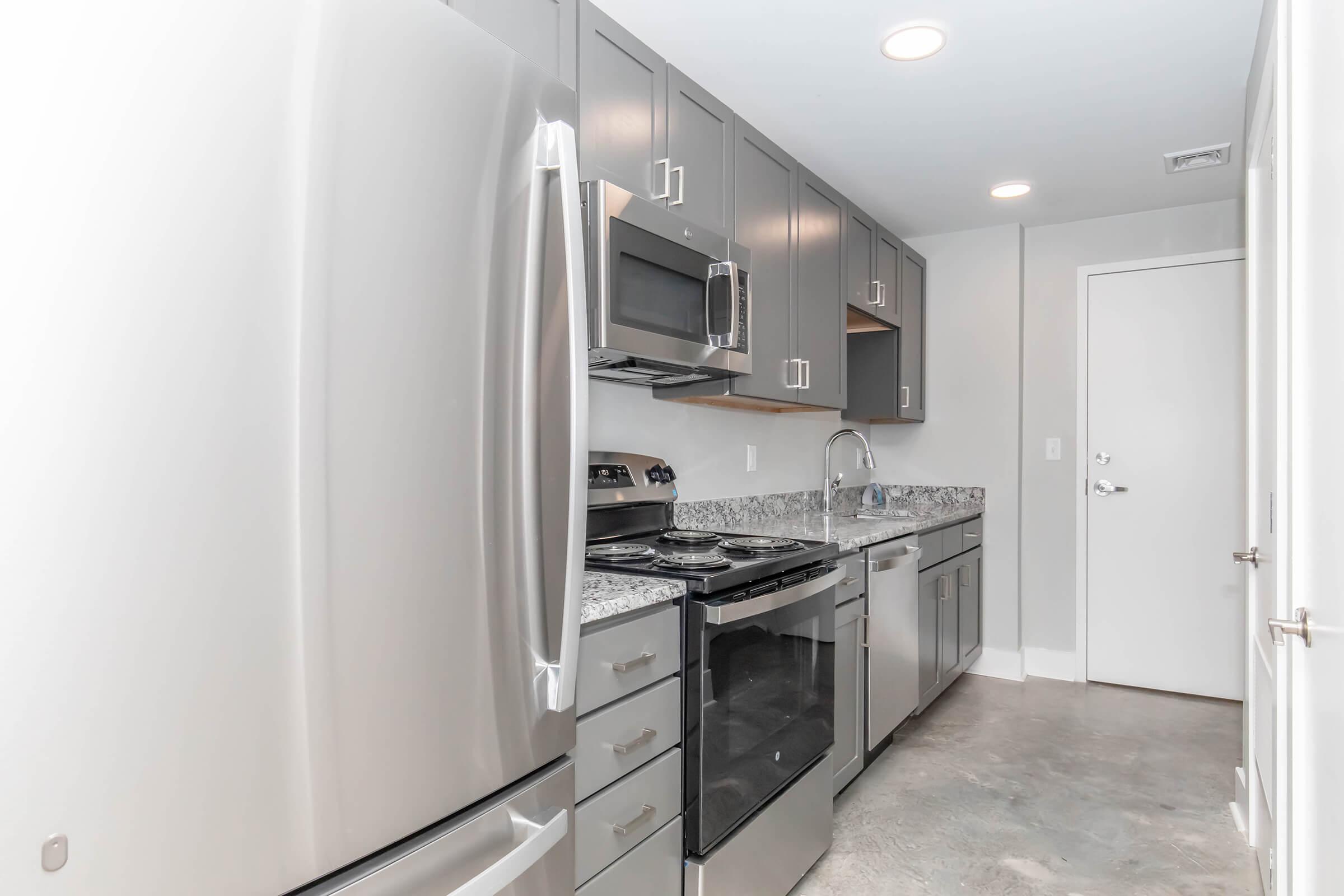 Modern kitchen featuring stainless steel appliances, including a refrigerator, oven, microwave, and dishwasher. The countertop is made of granite, and the cabinets are sleek and gray. A single-basin sink is installed, and the space has a clean, contemporary look with neutral-colored walls and concrete flooring.