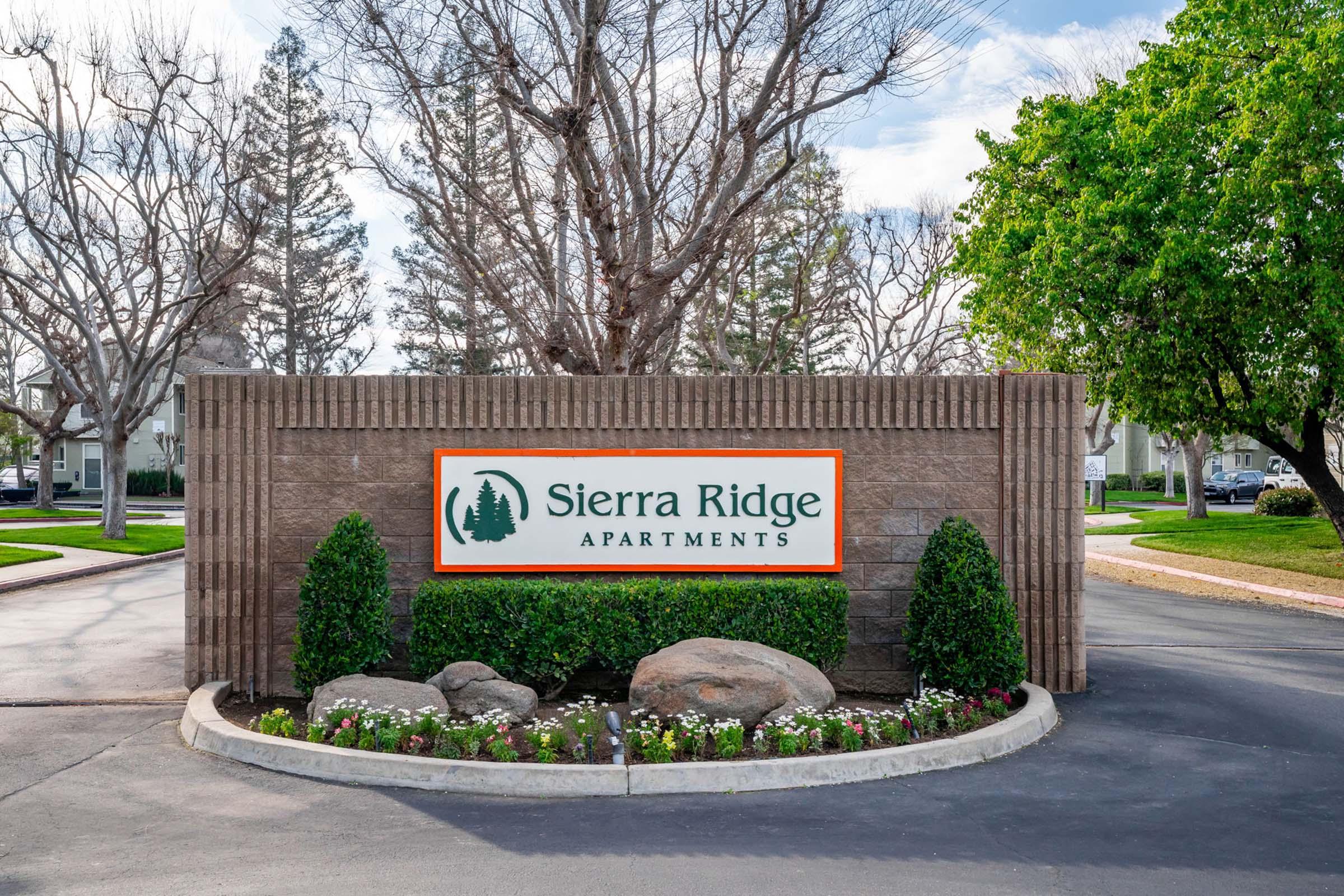 Sign for Sierra Ridge Apartments, featuring the name prominently displayed. Surrounded by landscaped greenery, including small trees and flower beds. The scene is bright, with blue sky and clouds in the background. The entrance is well-maintained, indicating a welcoming atmosphere.