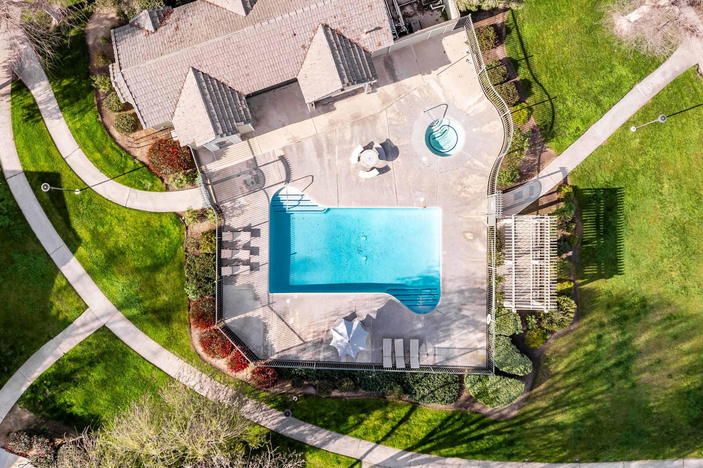 An aerial view of a residential backyard featuring a clear swimming pool, surrounded by a patio area. Nearby, there are lounge chairs, a shaded pergola, and a landscaped lawn with pathways leading to various garden areas. The scene is bright and sunny, showcasing outdoor relaxation space.