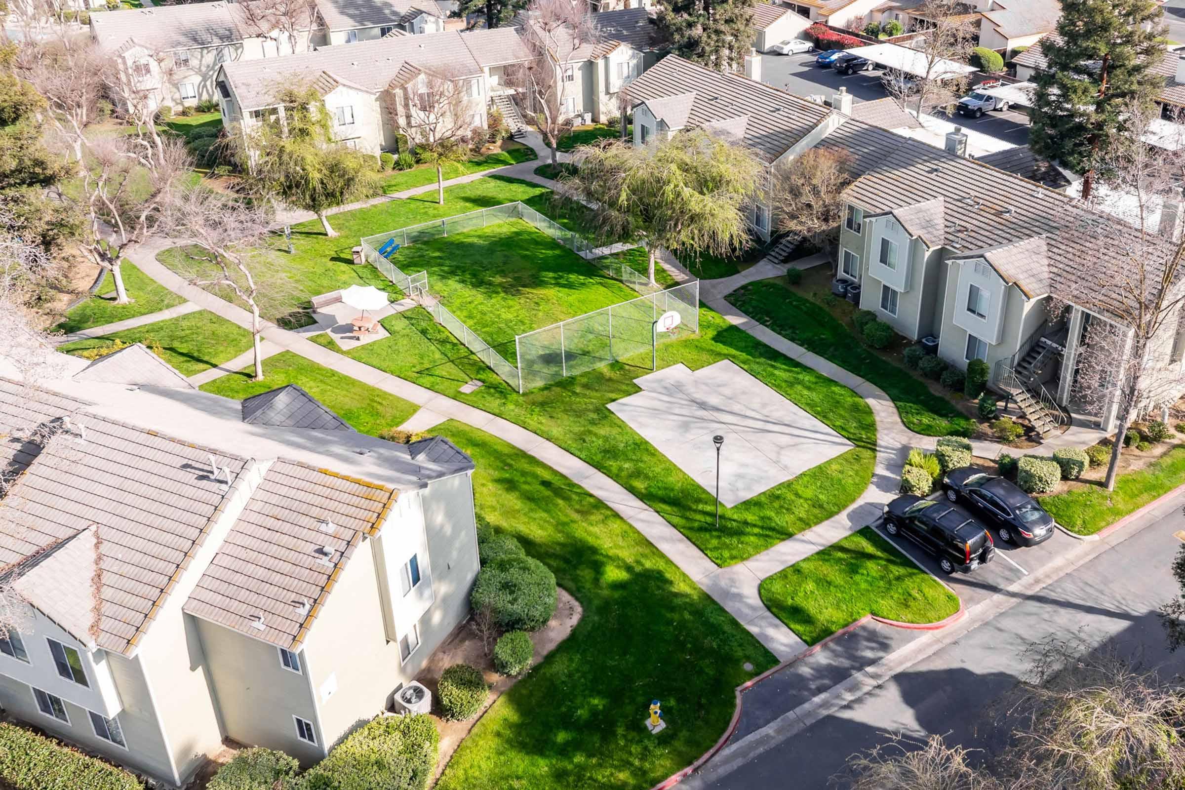 Aerial view of a residential area featuring several two-story apartment buildings surrounded by well-maintained green lawns, a basketball court, and a small playground. Pathways connect the buildings, providing access to outdoor spaces. Trees are scattered throughout the area.