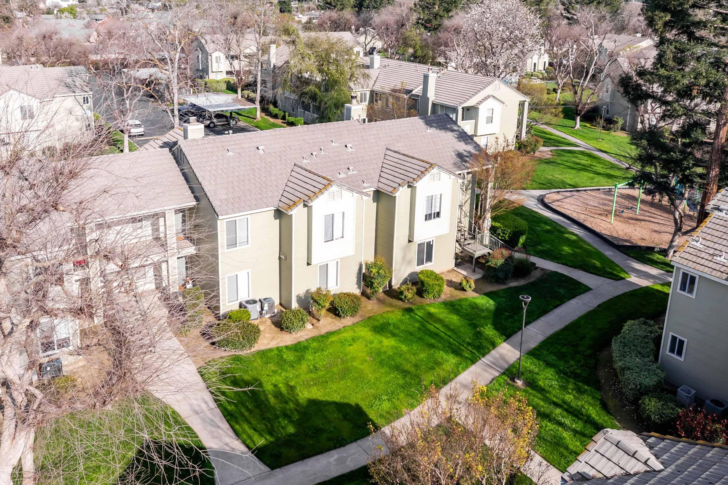 Aerial view of a residential complex featuring several two-story buildings surrounded by green lawns and walking pathways. Trees are visible in the background, and a playground can be seen nearby, indicating a family-friendly environment.
