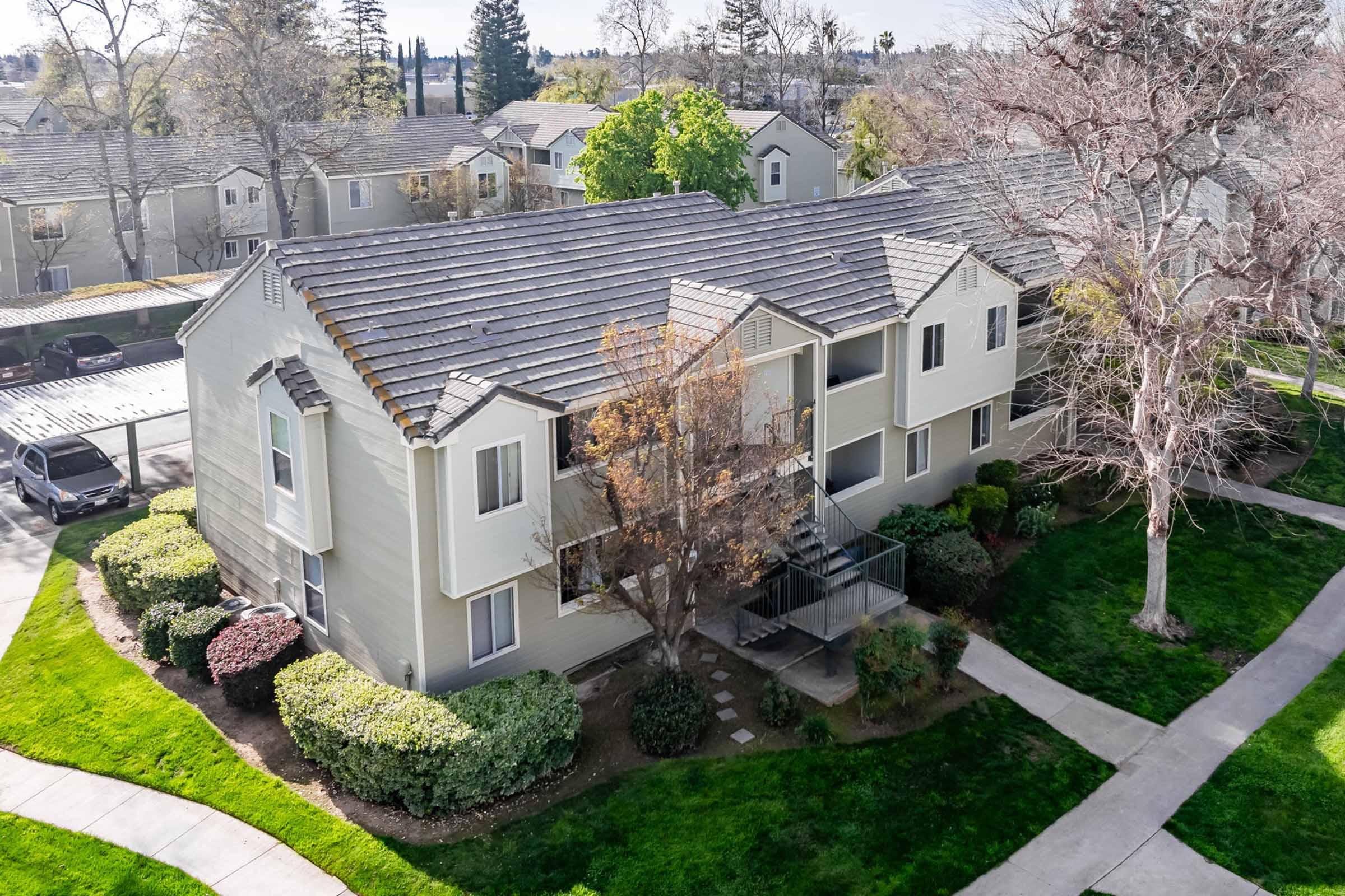 Aerial view of a multi-unit residential building surrounded by well-maintained landscaping, including shrubs and trees. The building features a light-colored exterior, two levels, and balconies. Sidewalks lead through the green lawn area. Nearby parking spaces are visible.