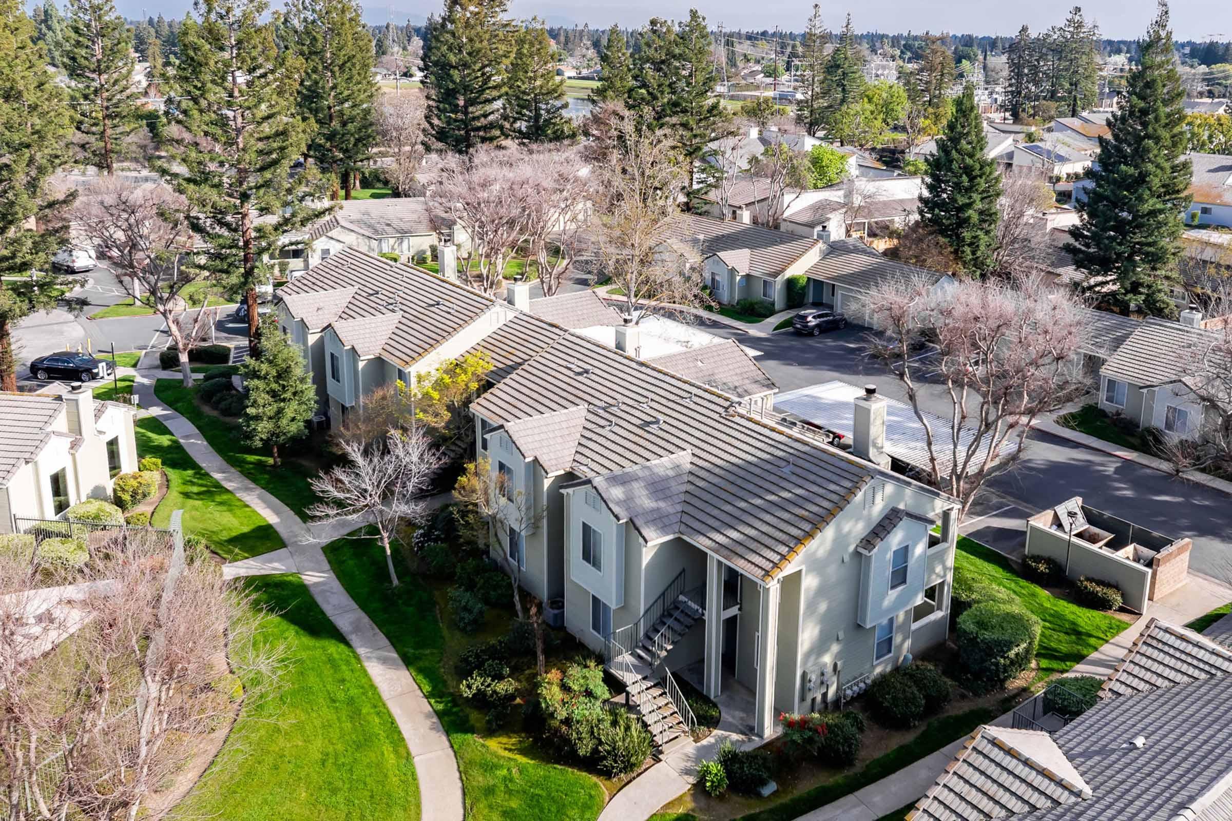 An aerial view of a residential neighborhood featuring multiple low-rise apartment buildings surrounded by well-maintained green lawns and trees. The scene shows walking paths, parking areas, and a mix of deciduous and evergreen trees, indicating a suburban setting.