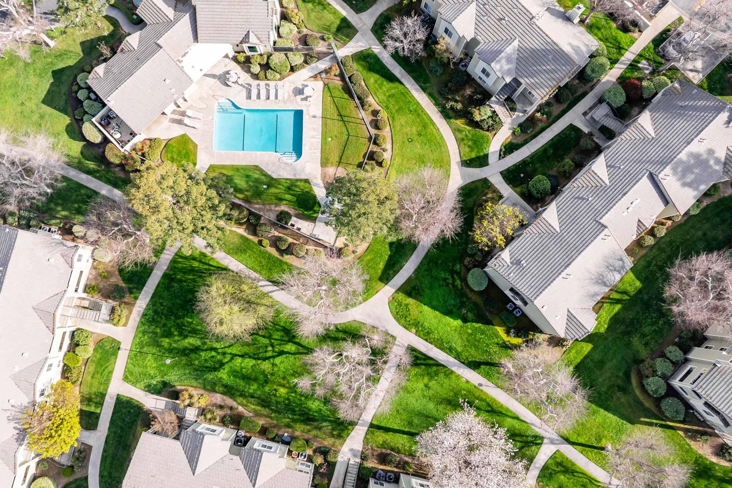 Aerial view of a suburban neighborhood featuring houses with well-maintained lawns and landscaping. A swimming pool is centrally located, surrounded by pathways and trees. The layout shows a mix of green spaces and residential structures, highlighting the organized community design.