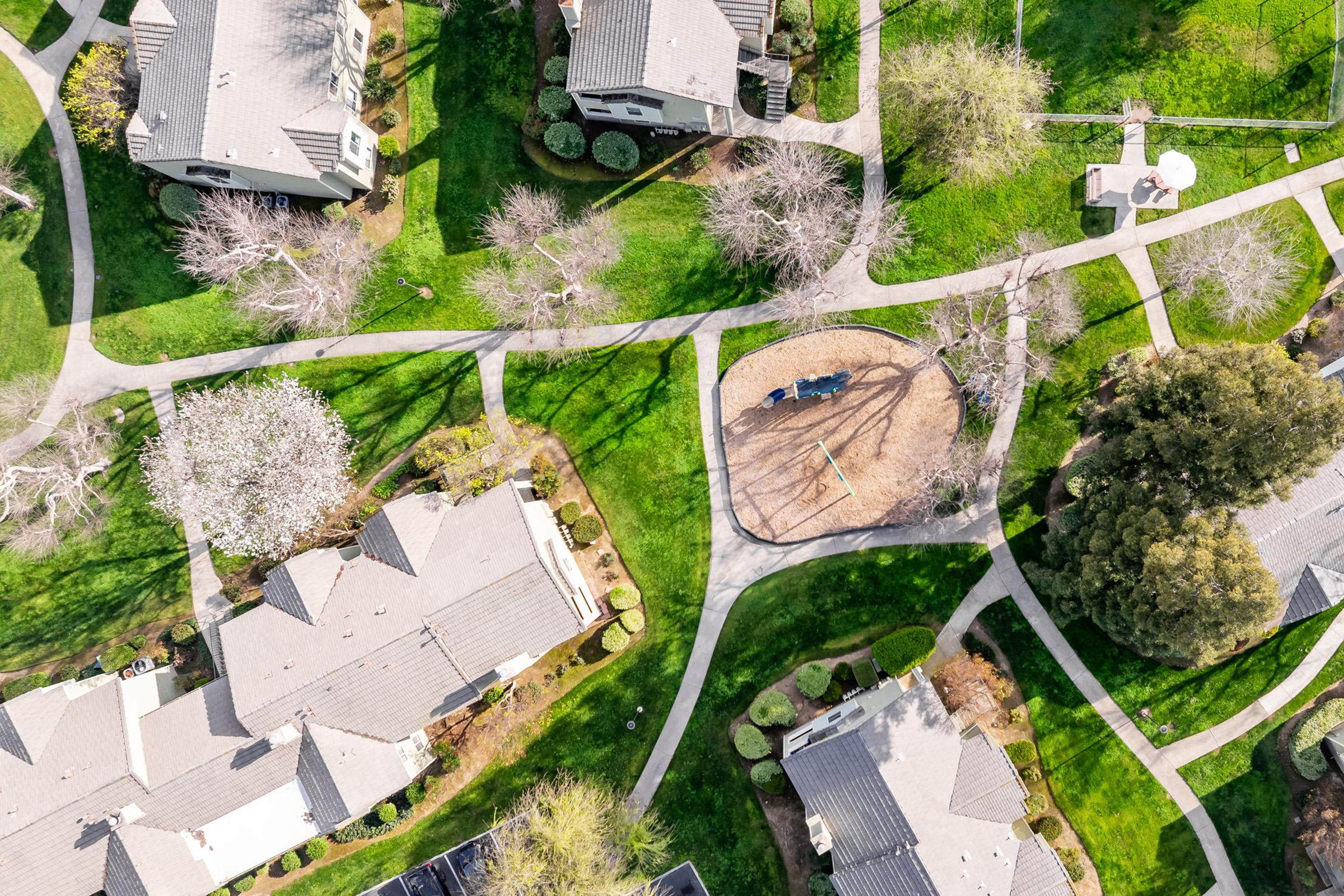 Aerial view of a residential neighborhood featuring several houses with gray roofs, manicured lawns, and walkways. A park area includes a sandbox and playground equipment, surrounded by trees. The layout shows a mix of winding paths and green spaces.