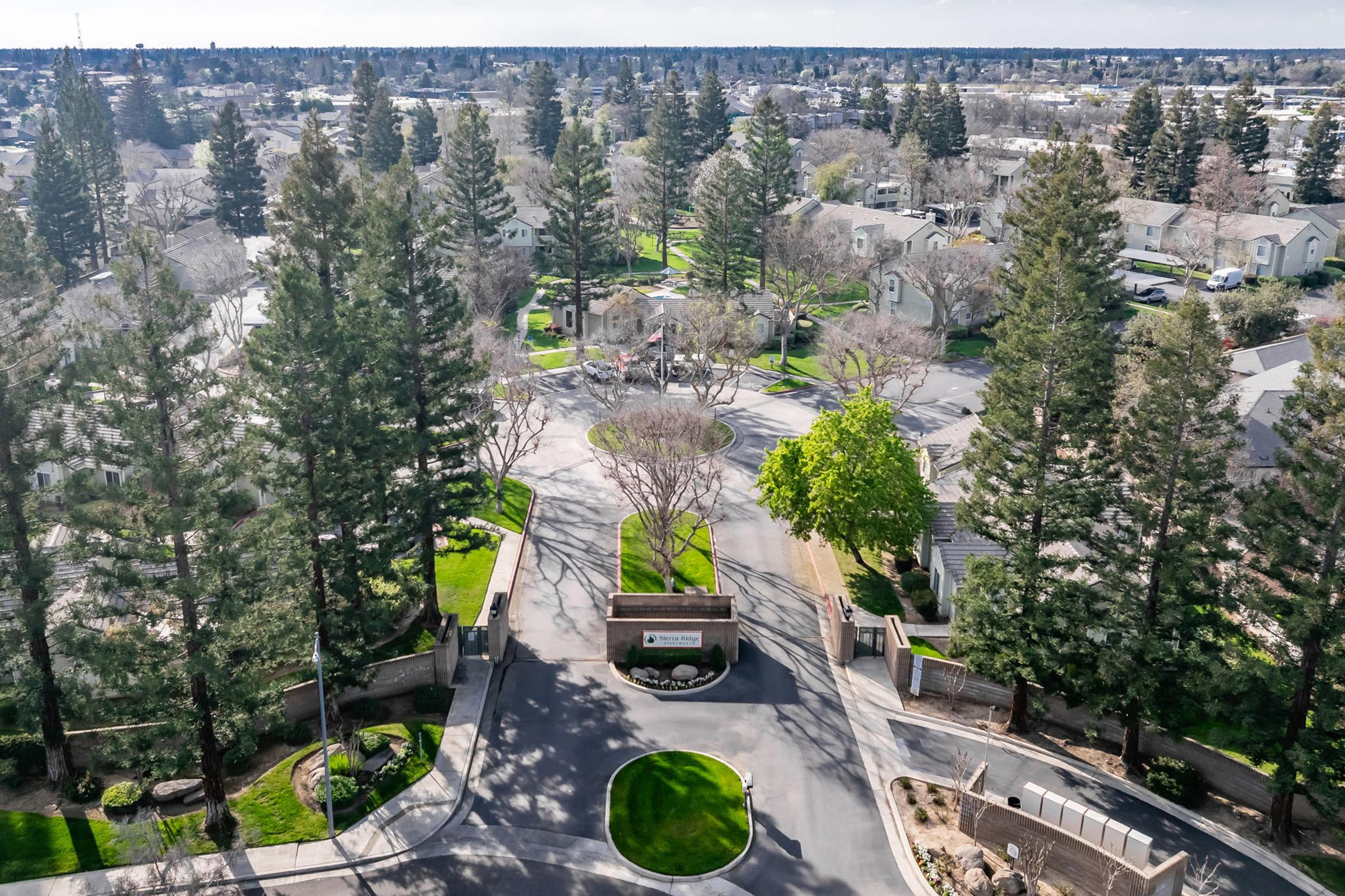 Aerial view of a residential community, showcasing tree-lined streets and well-maintained landscaping. A circular driveway is visible at the entrance, with a sign indicating the community's name. Surrounding greenery includes tall trees and manicured lawns, with houses and apartments visible in the background.