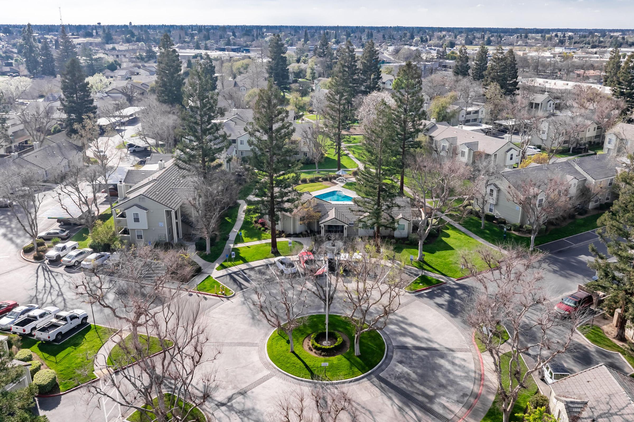 Aerial view of a residential neighborhood featuring several houses and a roundabout. Lush green lawns and trees surround the area, with a central landscaped circle. A swimming pool is visible, and cars are parked along the streets. The sky is clear, showcasing a peaceful suburban atmosphere.