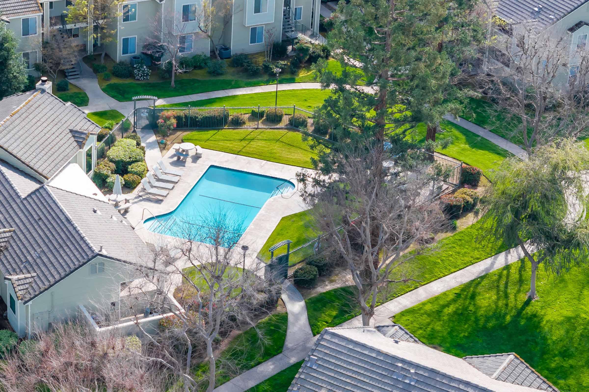 Aerial view of a residential complex featuring a swimming pool surrounded by green lawns and pathways. Several houses with roofs are visible, along with trees and landscaped areas, creating a serene outdoor environment.