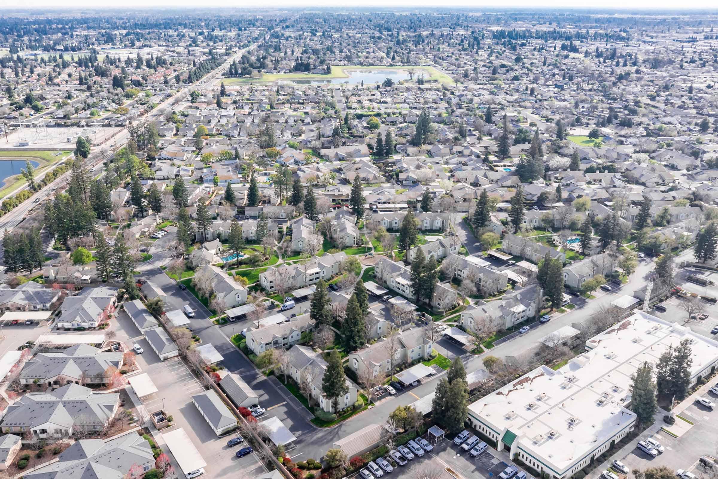 Aerial view of a suburban neighborhood featuring numerous homes with rooftops, tree-lined streets, and green spaces. Nearby, there are commercial buildings and a large parking lot. The landscape shows a mix of residential and commercial areas, with a distant view of a park or pond in the center.