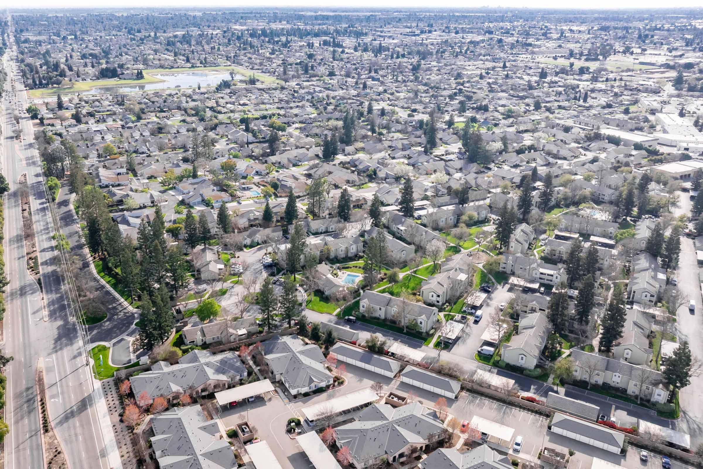 Aerial view of a densely populated suburban area featuring numerous residential buildings and homes. The landscape includes patches of green trees and grass, along with a road running along one side. In the distance, patches of water and additional housing can be seen, illustrating the vastness of the neighborhood.