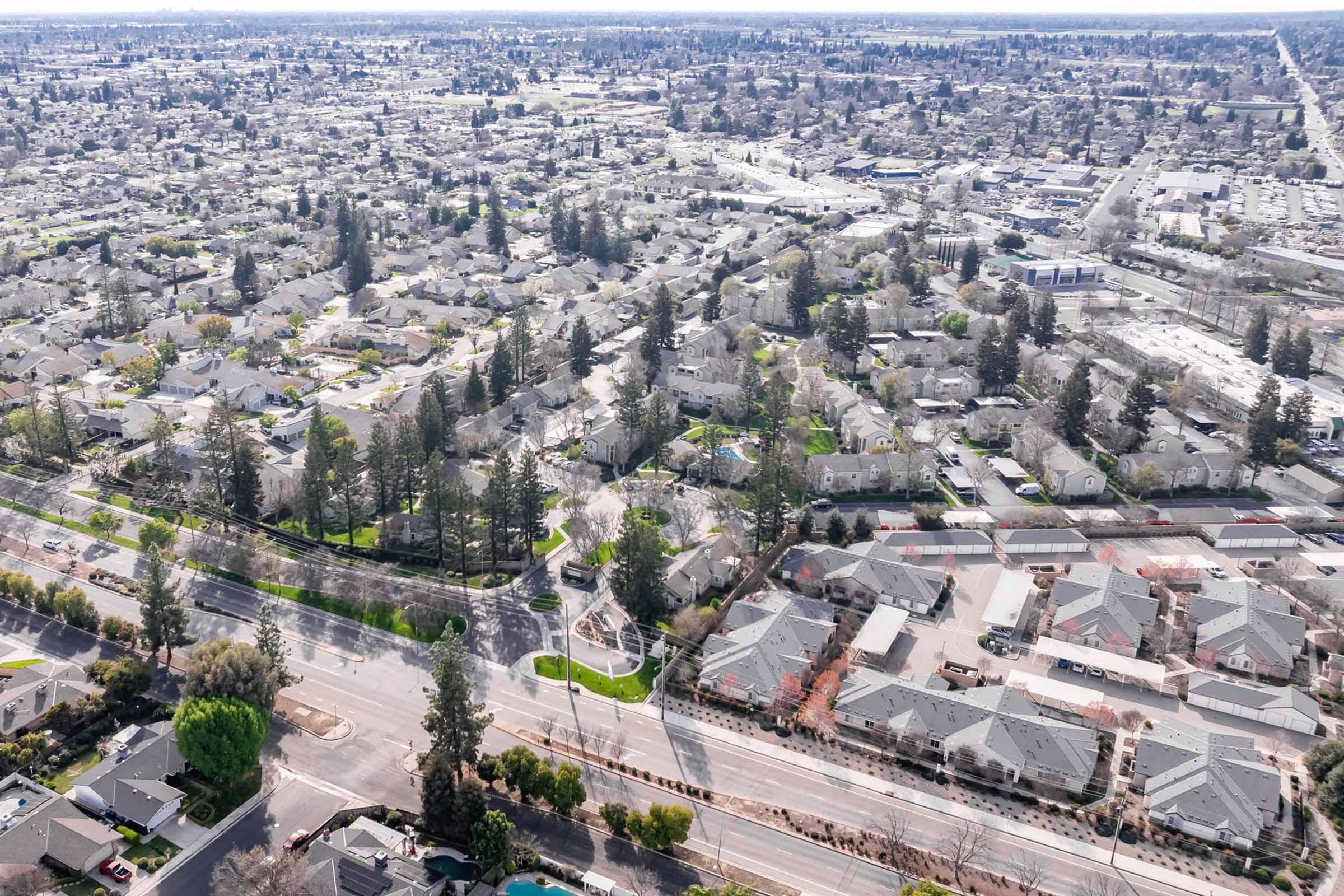 Aerial view of a suburban neighborhood featuring a dense arrangement of residential homes, streets, and green spaces. Tall trees line some areas, while visible roads and commercial buildings are in the background, highlighting the layout of the community. The landscape appears peaceful and well-maintained.