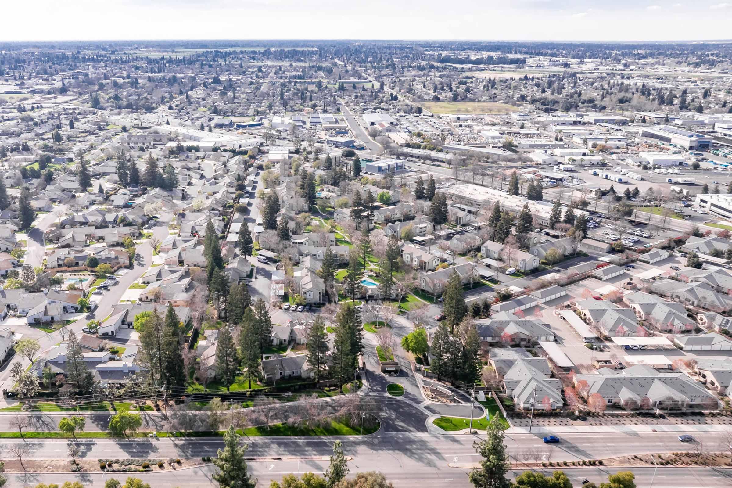 Aerial view of a suburban neighborhood featuring neatly arranged houses surrounded by trees, parks, and roads. In the distance, commercial areas and open spaces are visible, showcasing a blend of residential and urban development. Clear skies provide good visibility of the layout.
