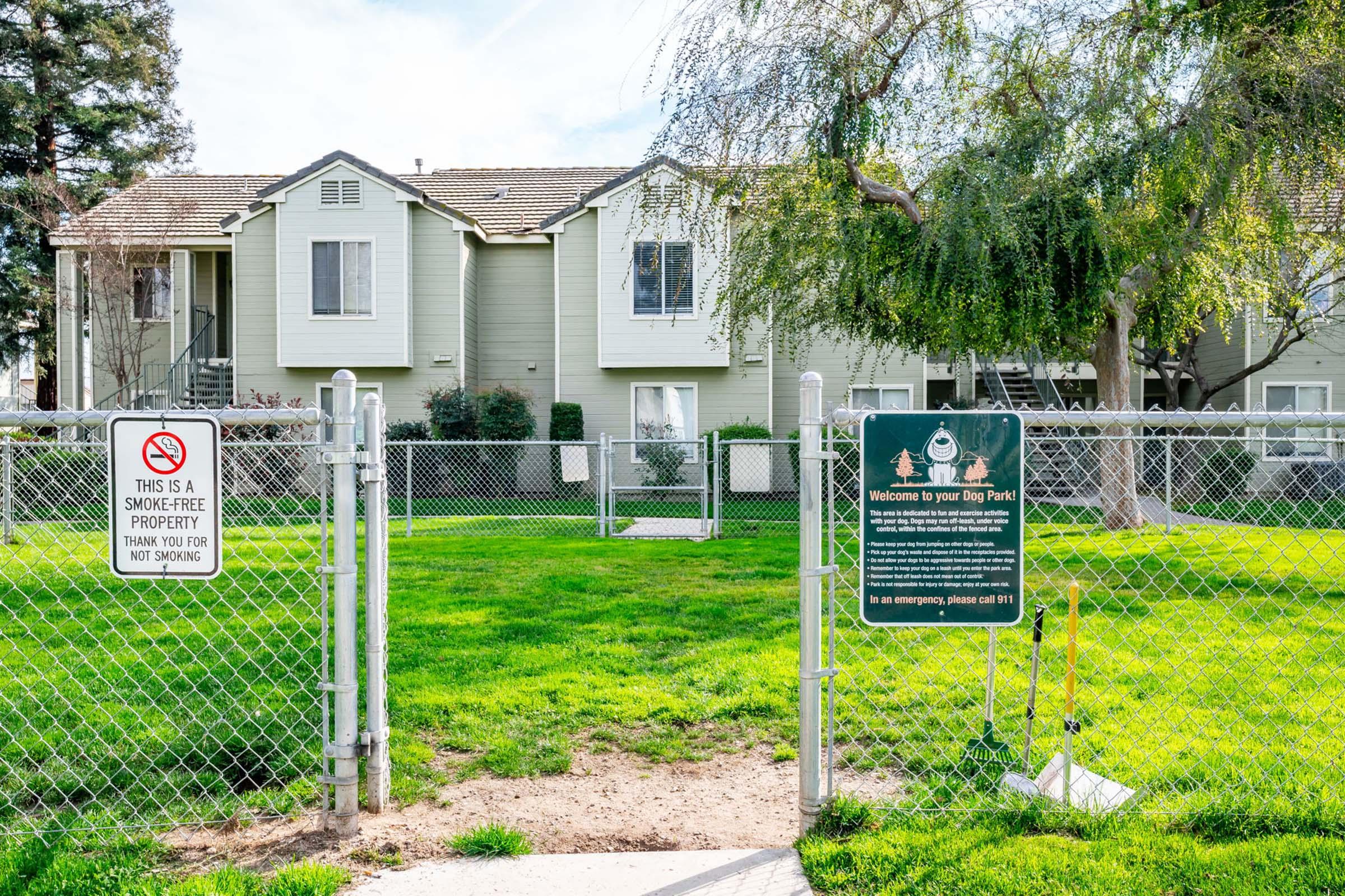 A fenced green area with a sign that reads "Welcome to your Dog Park." In the background, there are two-story residential buildings. A no smoking sign is visible near the entrance of the park. The scene has a well-maintained lawn and some trees.