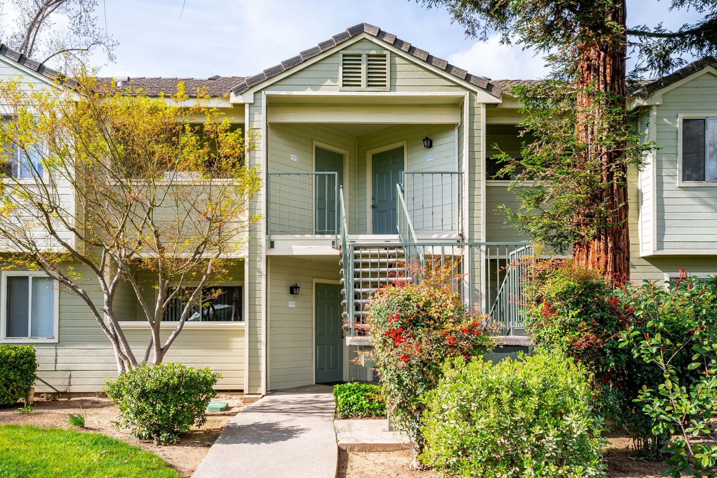 Two-story apartment building with a light green exterior. The entrance features a staircase leading to a second-floor door. Surrounding the building are well-maintained shrubs and trees, creating a landscaped area. A pathway leads to the entrance, inviting and well-lit.