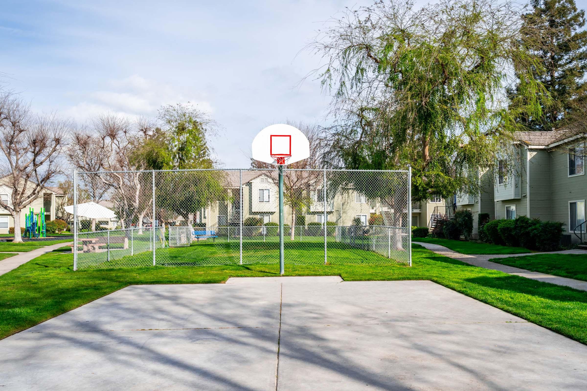 A basketball court surrounded by a chain-link fence, featuring a hoop with a red square backboard. In the background, there are residential buildings and green grass, with trees lining the area under a clear blue sky. The scene is well-lit, suggesting a sunny day.