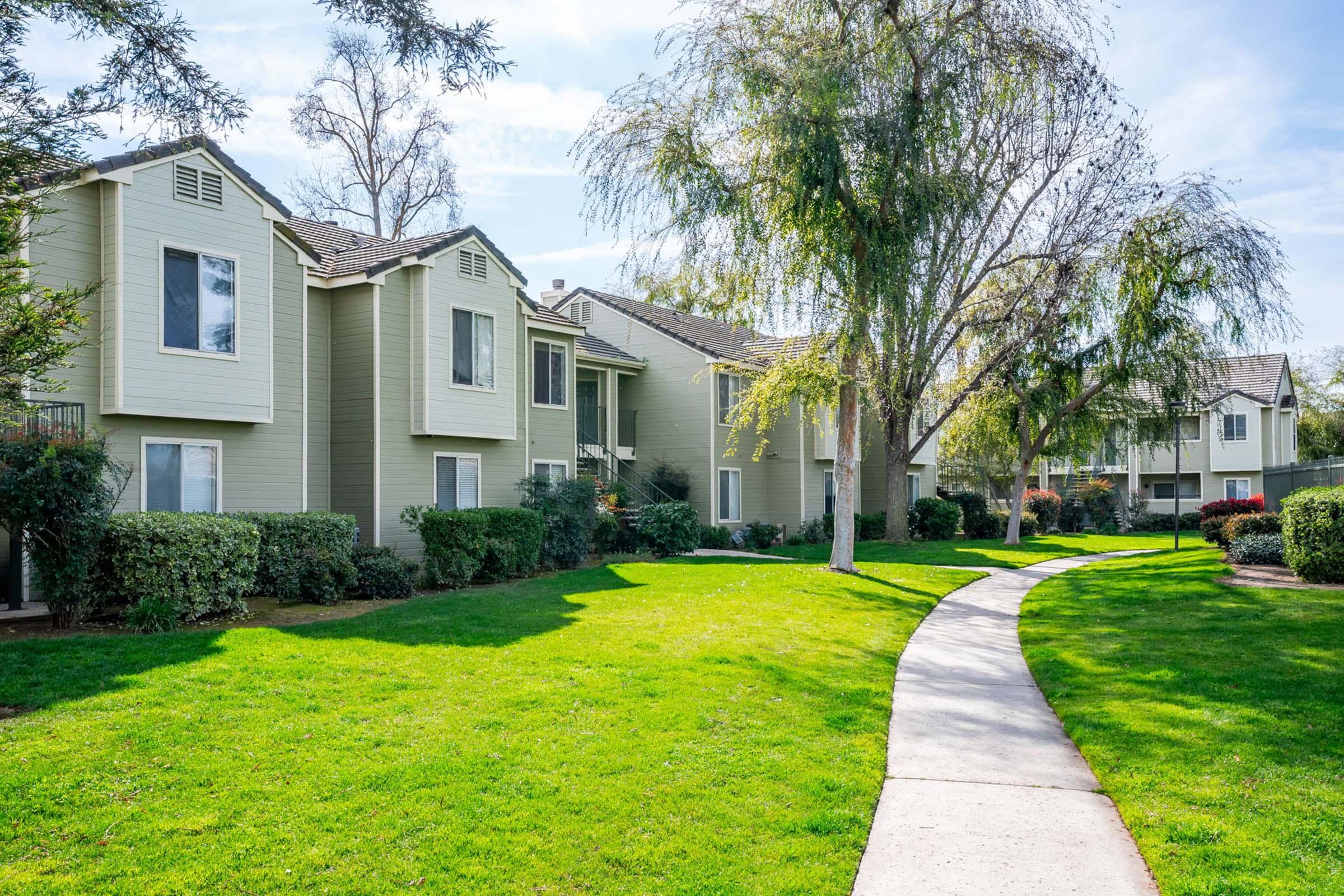 A well-maintained residential complex with two-story green apartment buildings surrounded by lush grass and shrubs. A concrete pathway winds through the landscaped area, leading past trees and greenery under a clear blue sky.