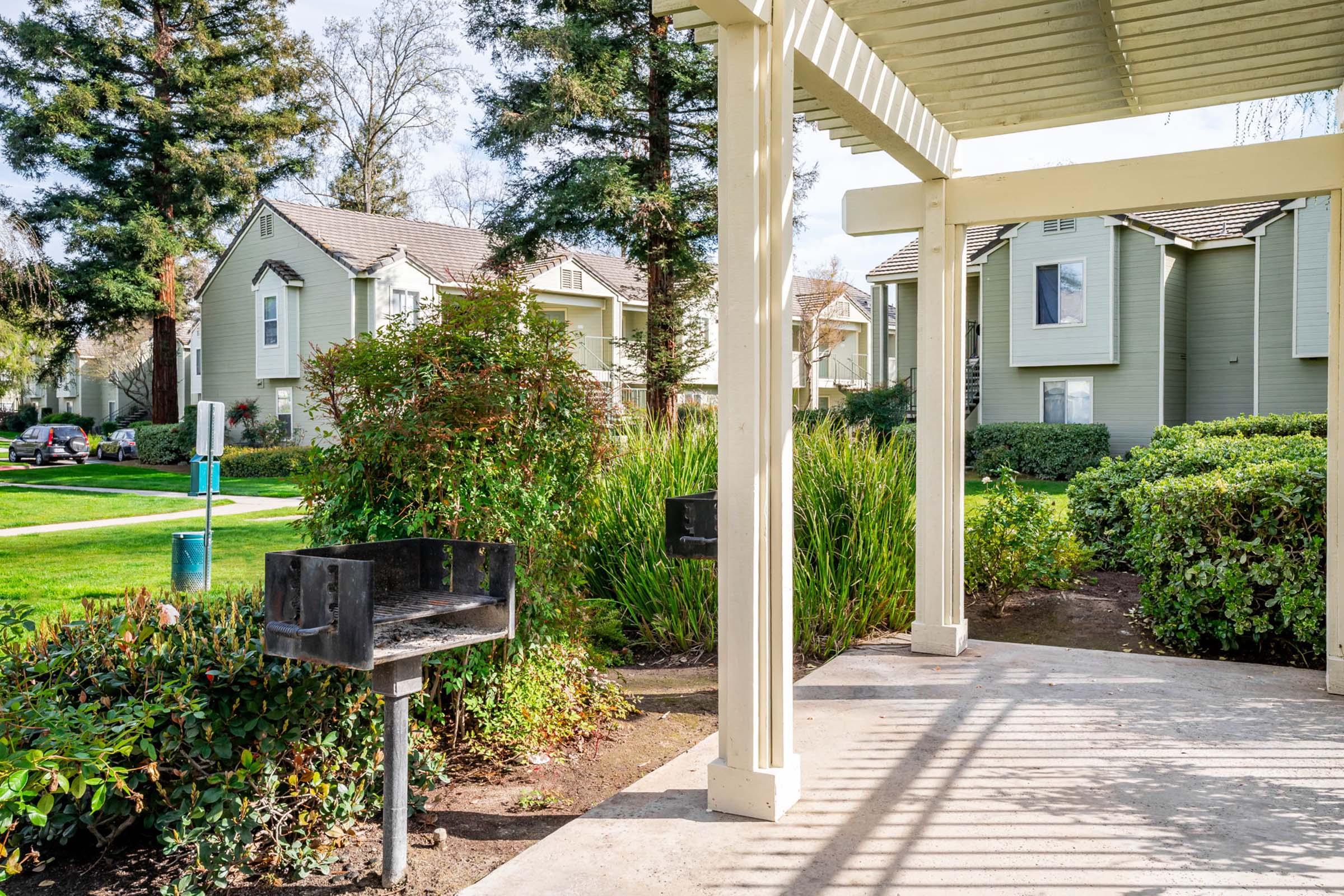 A view of a shaded outdoor space featuring a charcoal grill next to a walkway. Surrounding greenery includes bushes and trees, with residential buildings visible in the background. The scene is bright and inviting, suggesting a communal area for gatherings or barbecues.