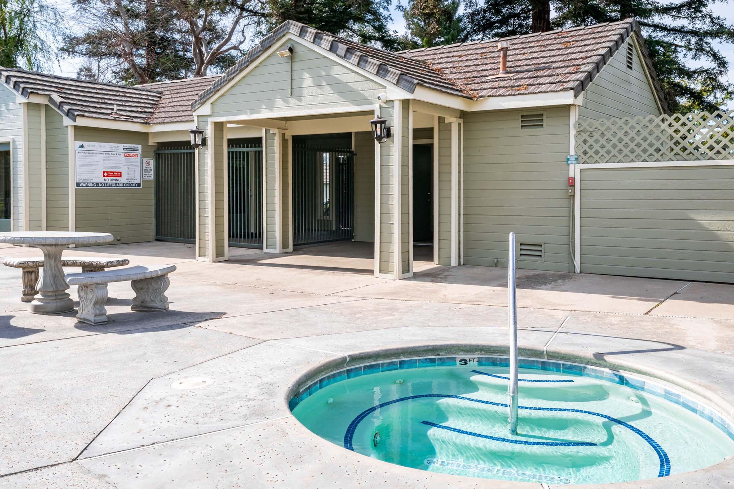 A well-maintained pool area featuring a hot tub with water jets, surrounded by stone seating and a wooden fence. The backdrop includes a light green building with multiple entrances and a sign. Lush trees provide a natural setting, enhancing the relaxing atmosphere.