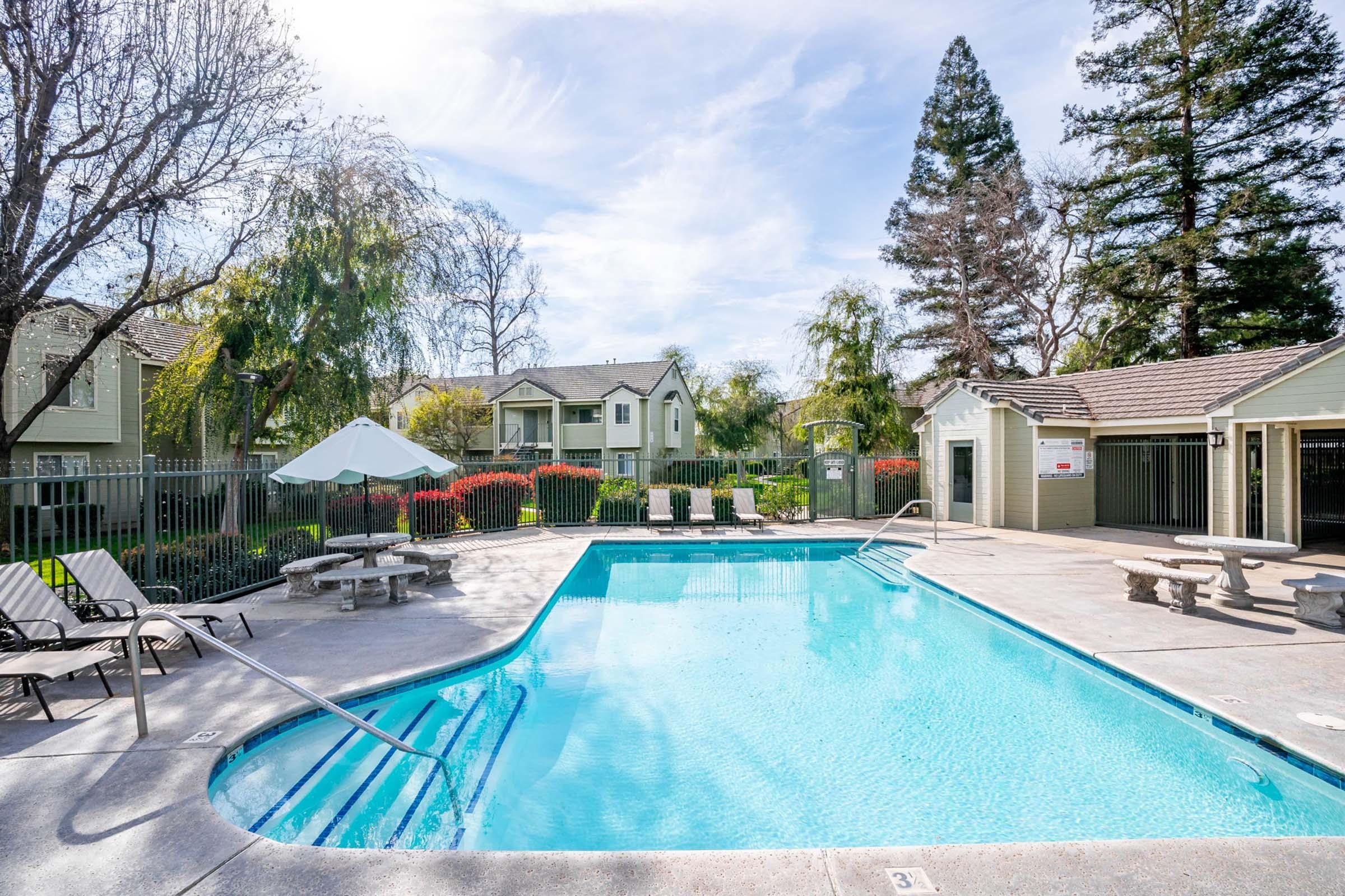 A tranquil outdoor swimming pool surrounded by lounge chairs, near a shaded area and lush greenery. In the background, residential buildings are visible, with trees and landscaping adding a serene touch. The sky is clear with a few clouds, creating a peaceful atmosphere.