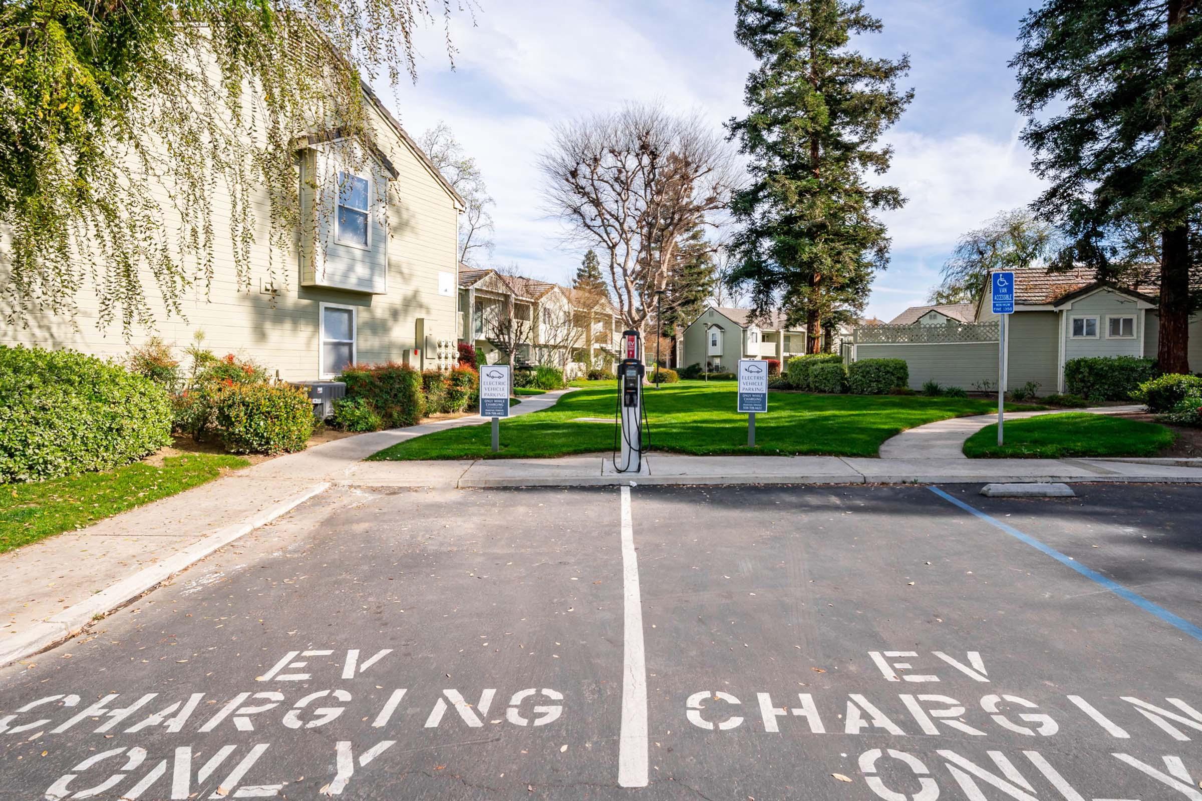 Two EV charging stations marked with "EV CHARGING ONLY" in a parking lot, surrounded by residential buildings and greenery. Sidewalks meander through the area, with a clear blue sky overhead.