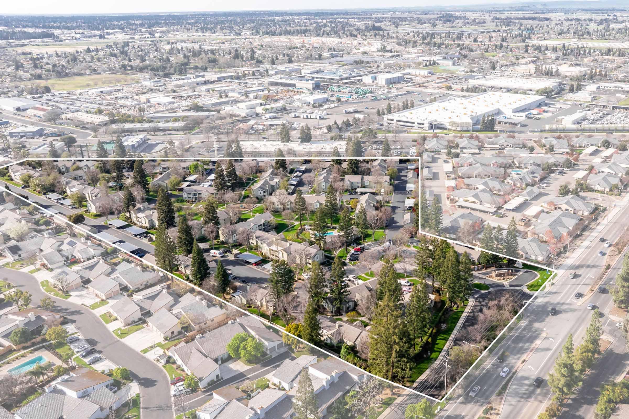 Aerial view of a suburban neighborhood with green parks and trees, surrounded by commercial areas and residential buildings. The image shows clearly defined property lines with street access, showcasing the layout of homes and landscaping in the area.