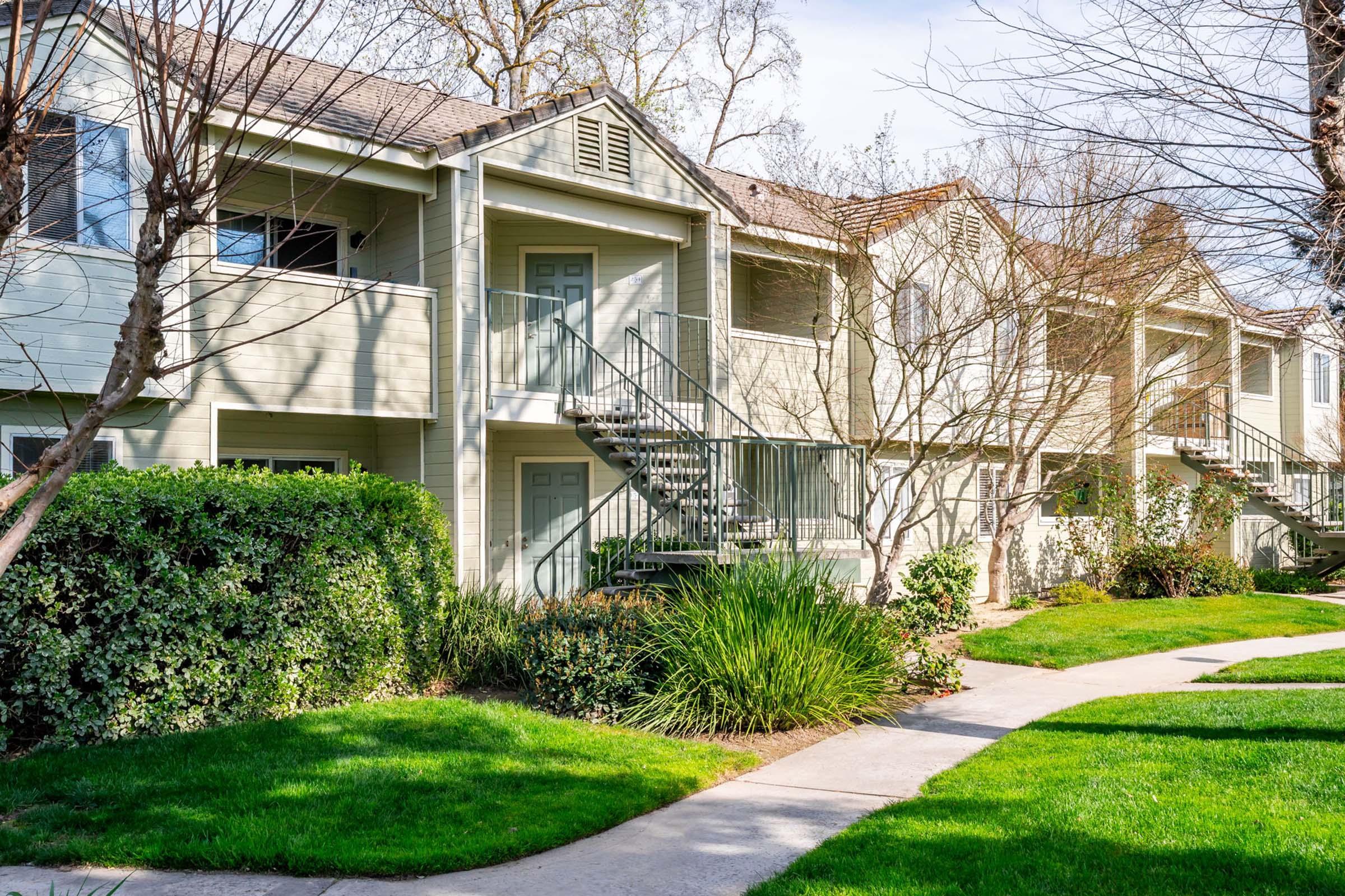 Two-story apartment building with a light green exterior and balconies, surrounded by neatly trimmed shrubs and grass. A paved walkway leads through the landscaping, with trees and a clear blue sky visible above.