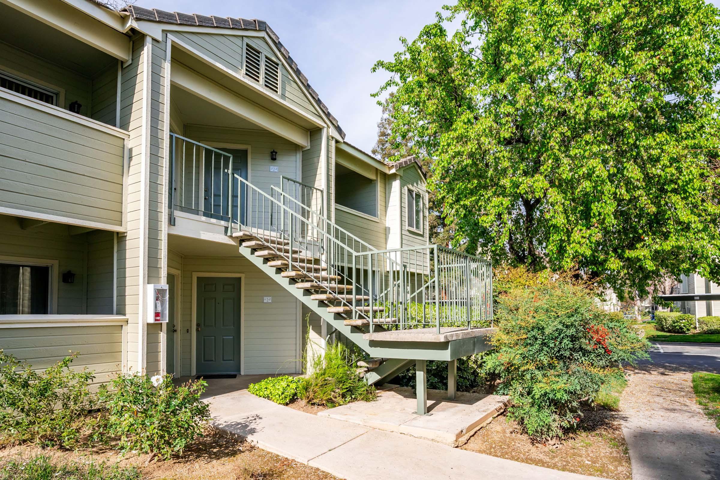 A two-story residential building with a green exterior. A set of stairs leads to the second floor, where a door is visible. Surrounding the building are landscaped plants and trees, providing a lush, green environment. The pathway in front is paved, leading towards the entrance.