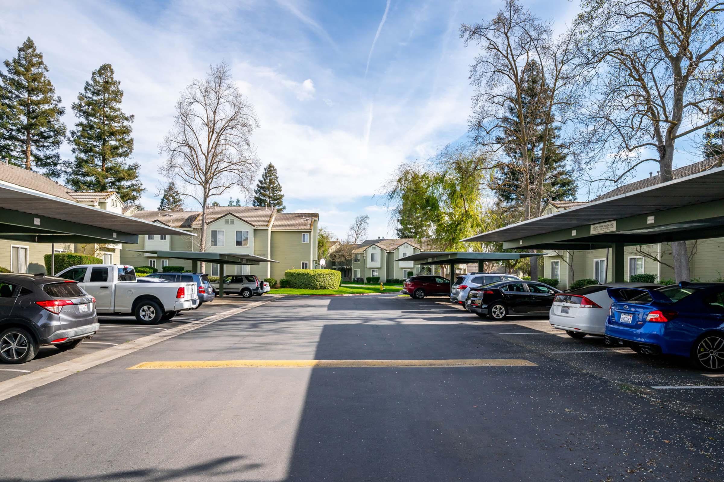 A view of a parking lot with several vehicles parked under covered spaces. In the background, there are residential buildings surrounded by trees and a blue sky with some clouds. The scene is bright and clear, depicting a suburban environment.