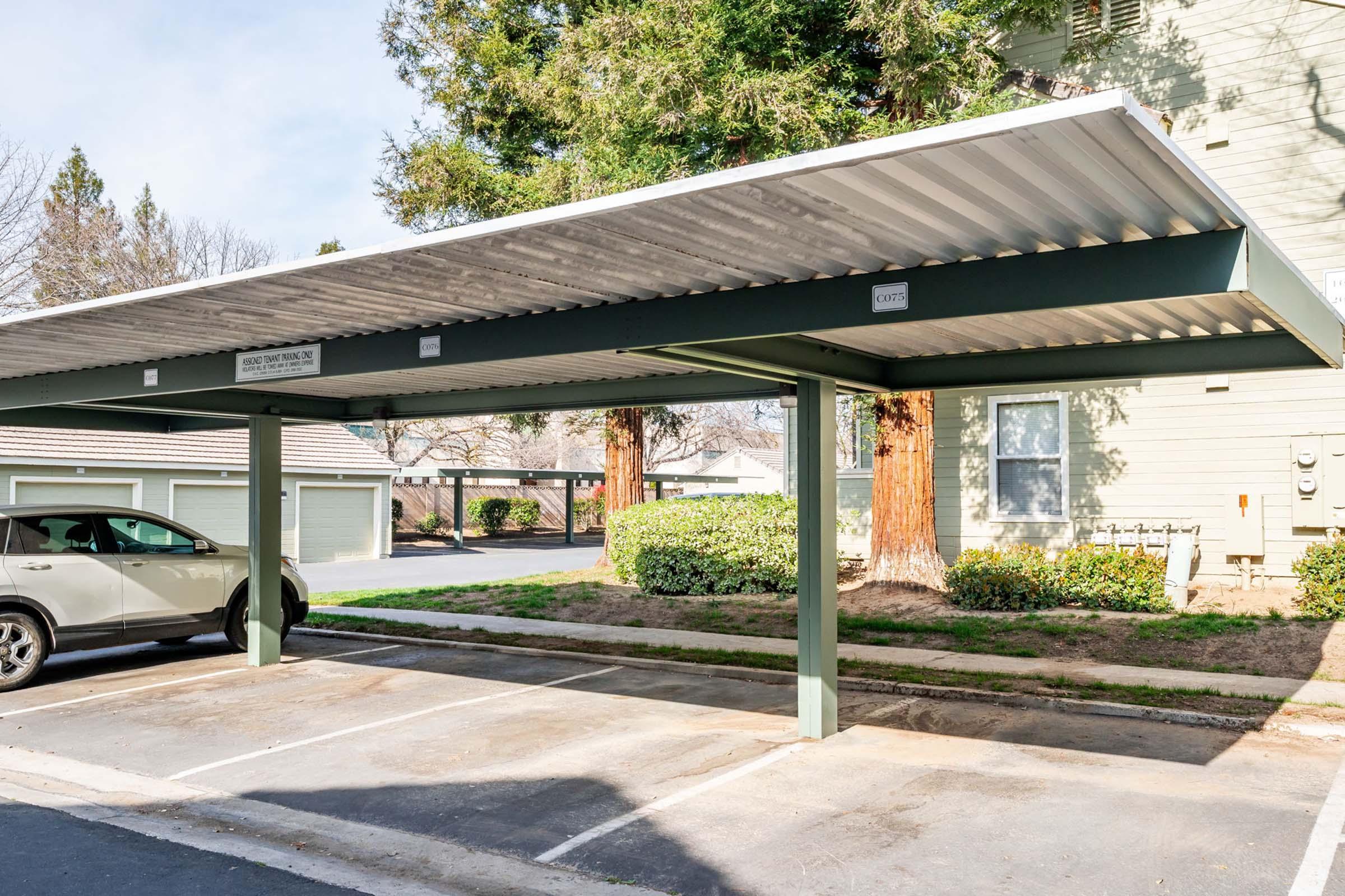 A covered parking space with a gray vehicle parked underneath. The structure features a metal roof supported by green columns. In the background, there are additional parking spots and a building with trees and shrubs nearby. The area appears to be well-maintained and is part of a residential complex.