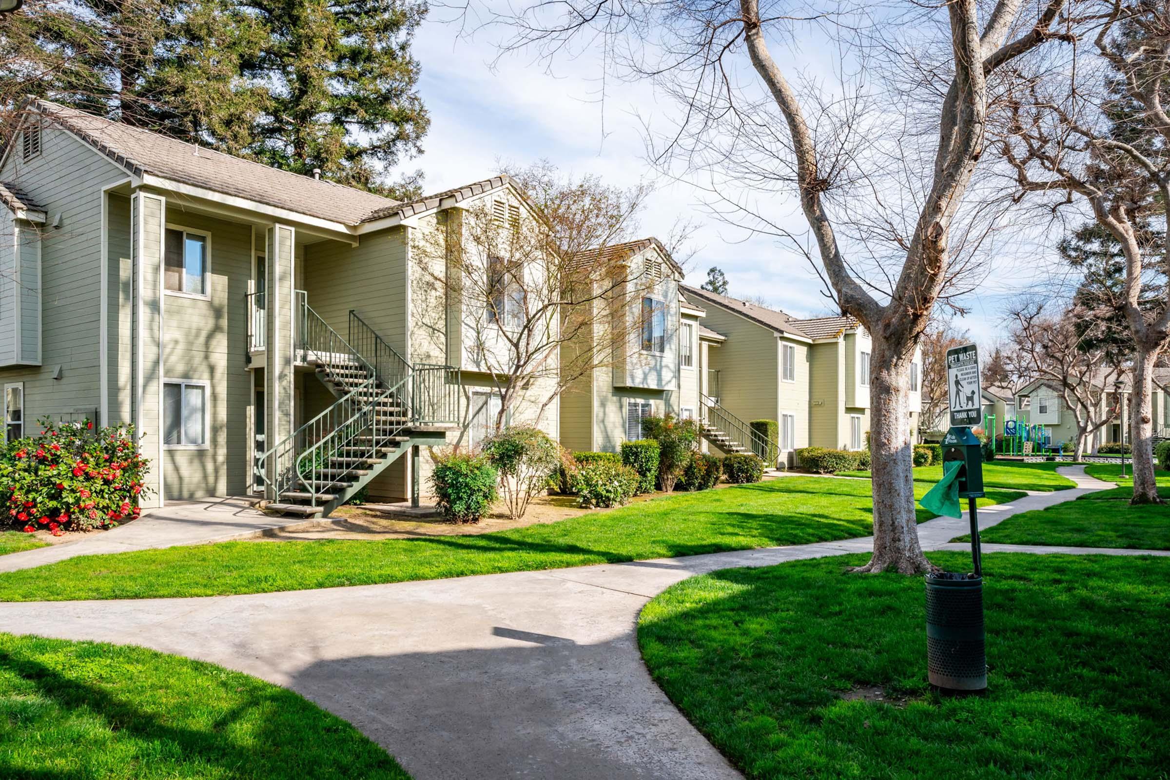 A brightly lit residential complex featuring two-story apartment buildings flanked by manicured lawns and small trees. A curved pathway winds through the greenery, leading to various entrances, while vibrant shrubs add color to the scene under a clear blue sky.