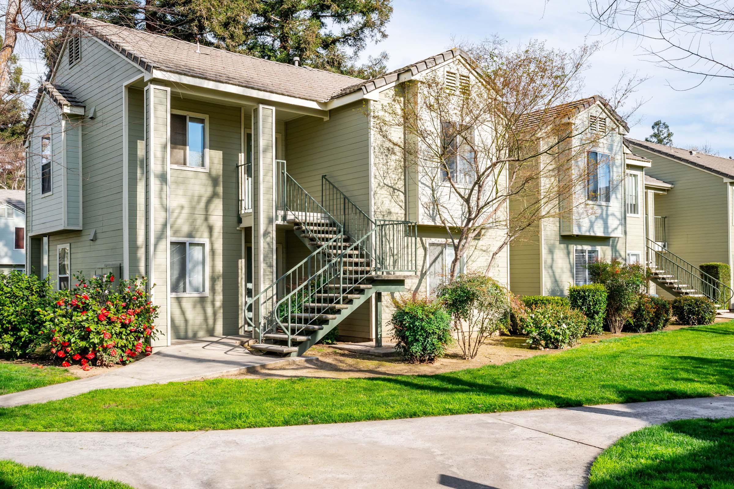Two multi-story apartment buildings with light green exteriors and balconies are surrounded by well-maintained lawns and shrubs. Steps lead up to the entrances, and a pathway runs along the front. Trees are visible in the background under a clear blue sky.