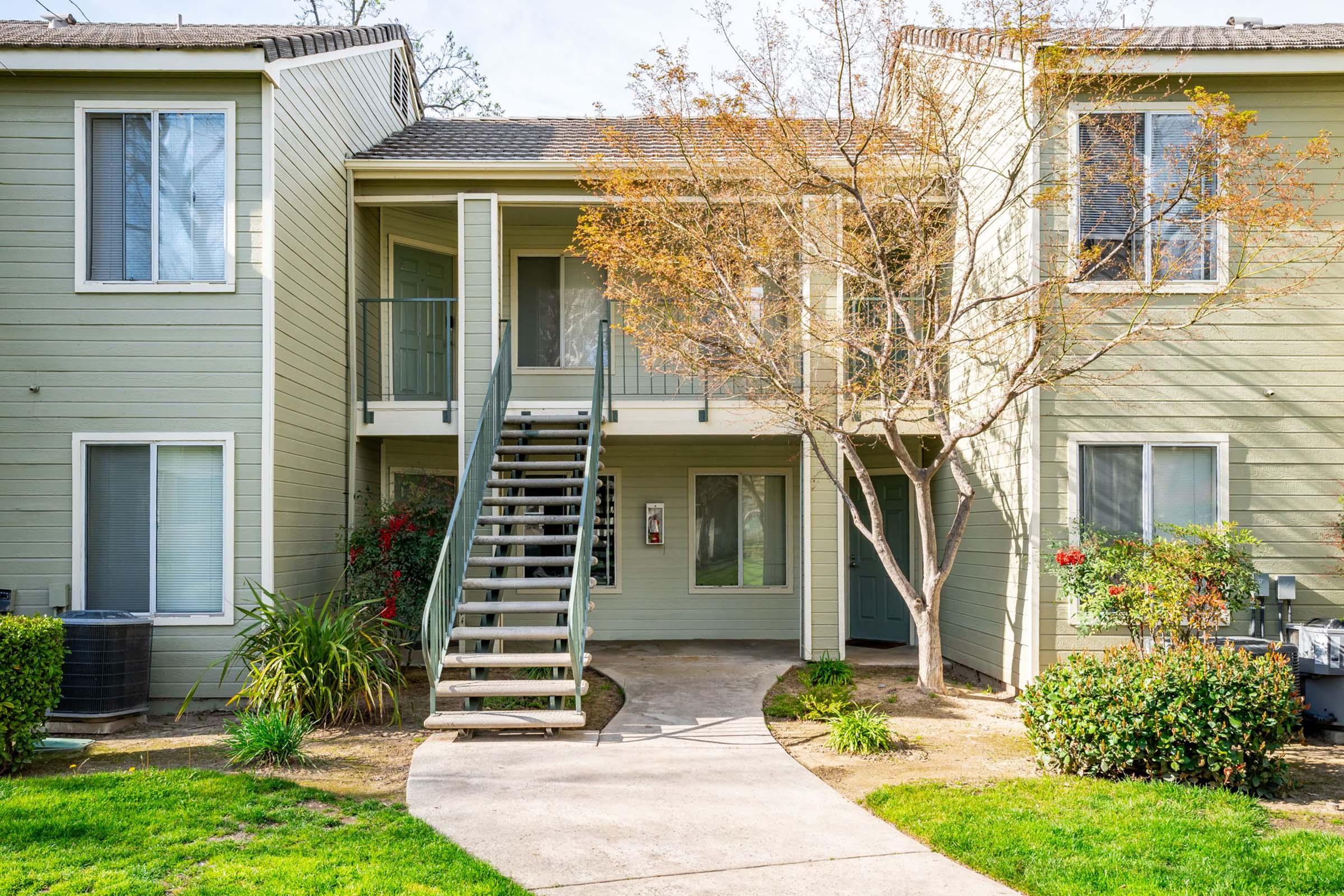 A view of a two-story apartment building with a curved pathway leading to the entrance. The building features light green siding, a staircase on the left, and several windows. There are shrubs and small trees surrounding the area, with a hint of autumn foliage. The setting is bright and welcoming.