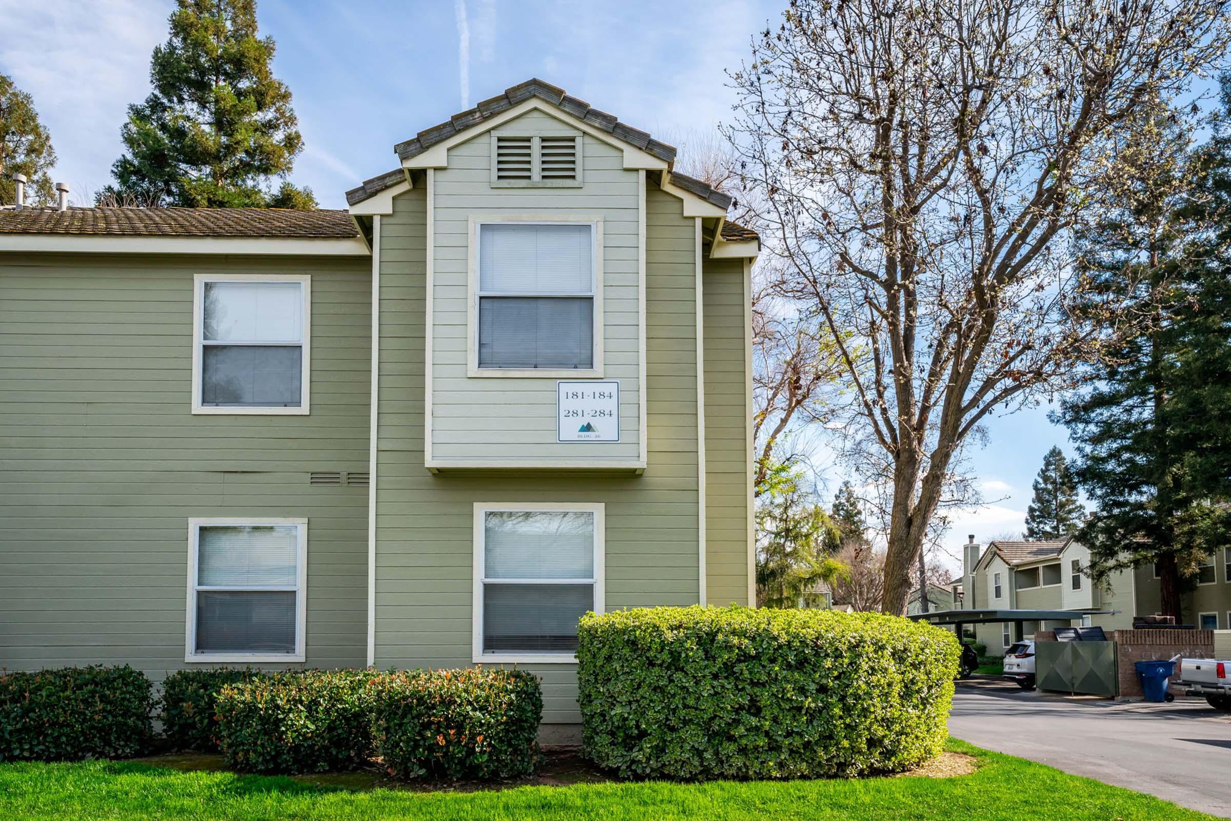 Two-story apartment building with a light green exterior. The upper floor features a centrally located window, flanked by two lower windows. A sign with numbers "1864" and "286" is visible on the upper window. Lush green shrubs line the base of the building, with trees in the background and a parking area visible to the side.