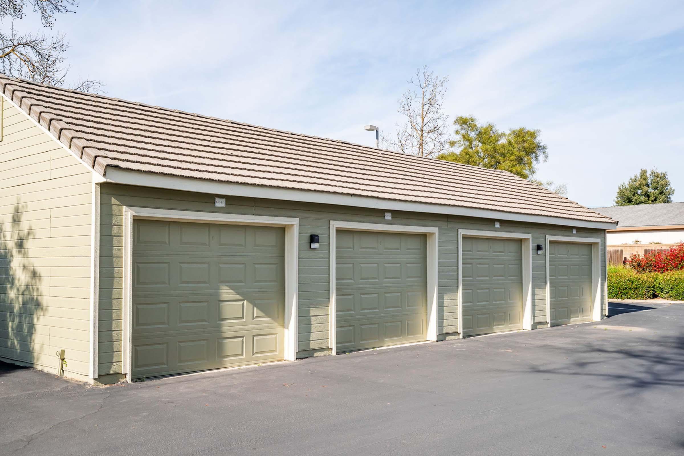 A row of three garages with green doors and a light brown exterior. The garages are set against a clear blue sky with a few trees in the background. The pavement in front is dark, and there are some shrubs and a building visible in the distance.