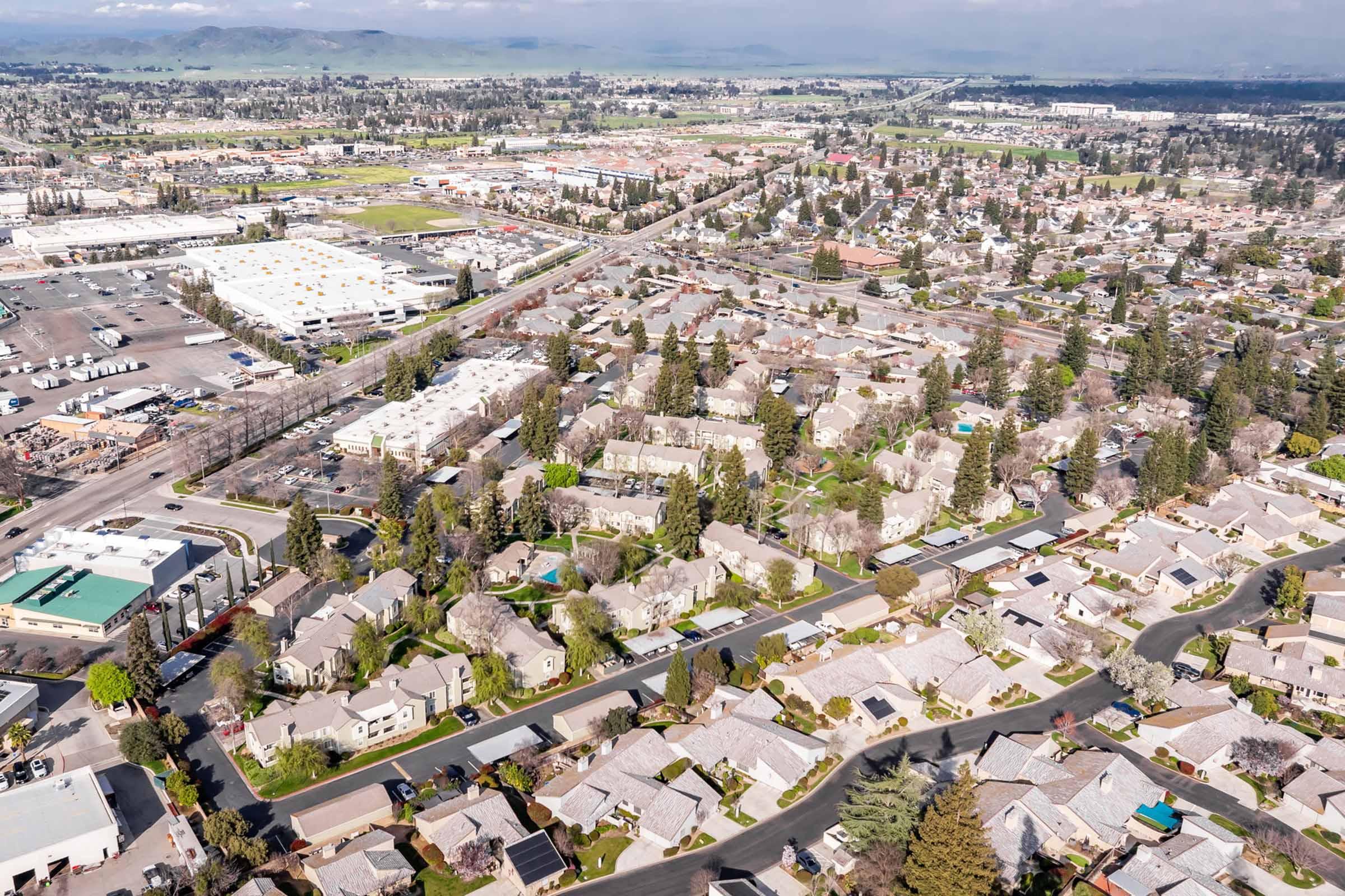 Aerial view of a suburban area showcasing residential neighborhoods with single-family homes, tree-lined streets, and nearby commercial zones. The landscape features a mix of greenery and developed areas, with mountains visible in the distance and a clear blue sky above.