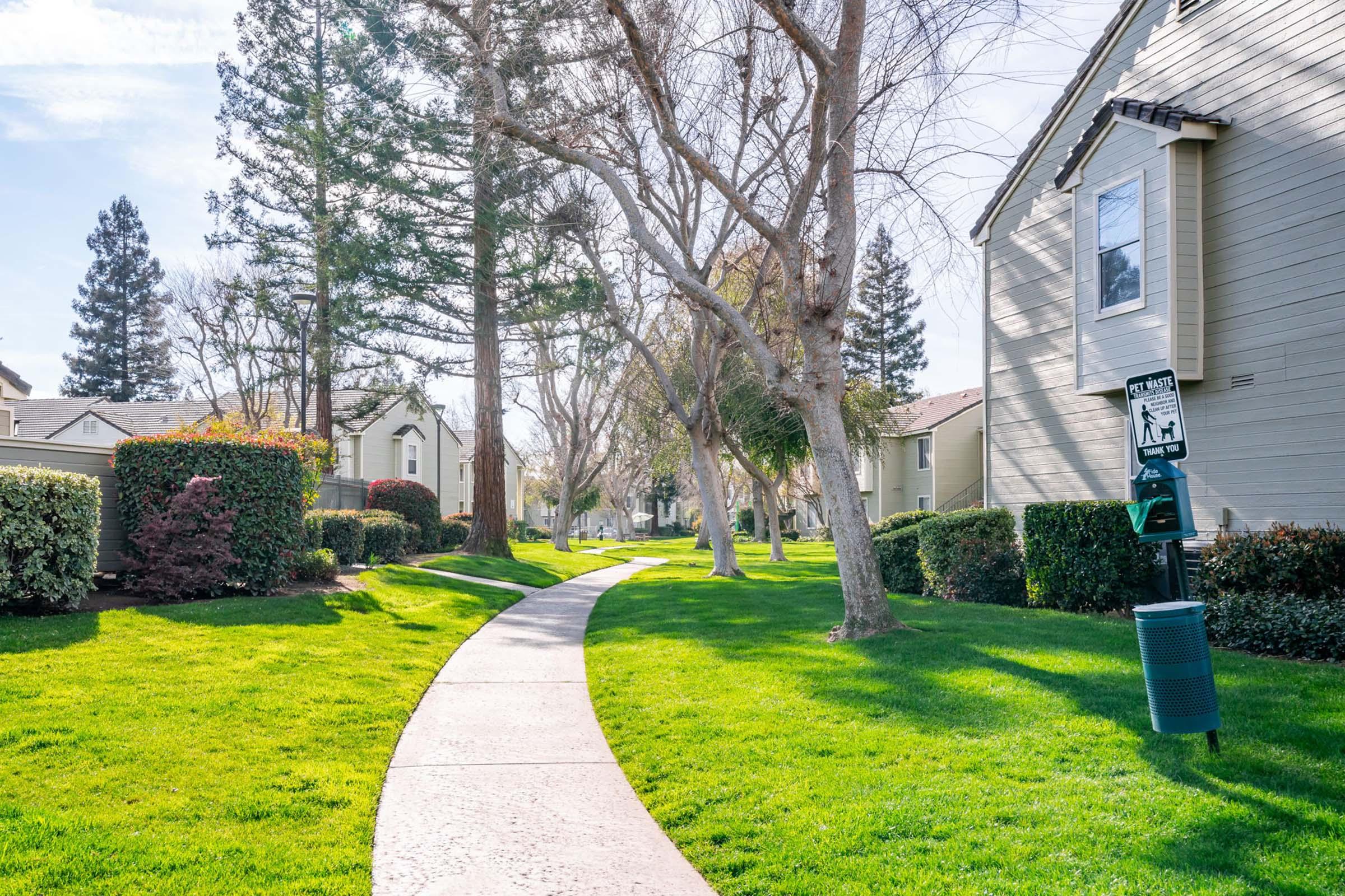 A peaceful pathway lined with trees and well-manicured grass, leading through a residential area. Houses are visible on either side, and a dog waste station is positioned along the path. The scene is bright and sunny, showcasing a serene suburban environment.