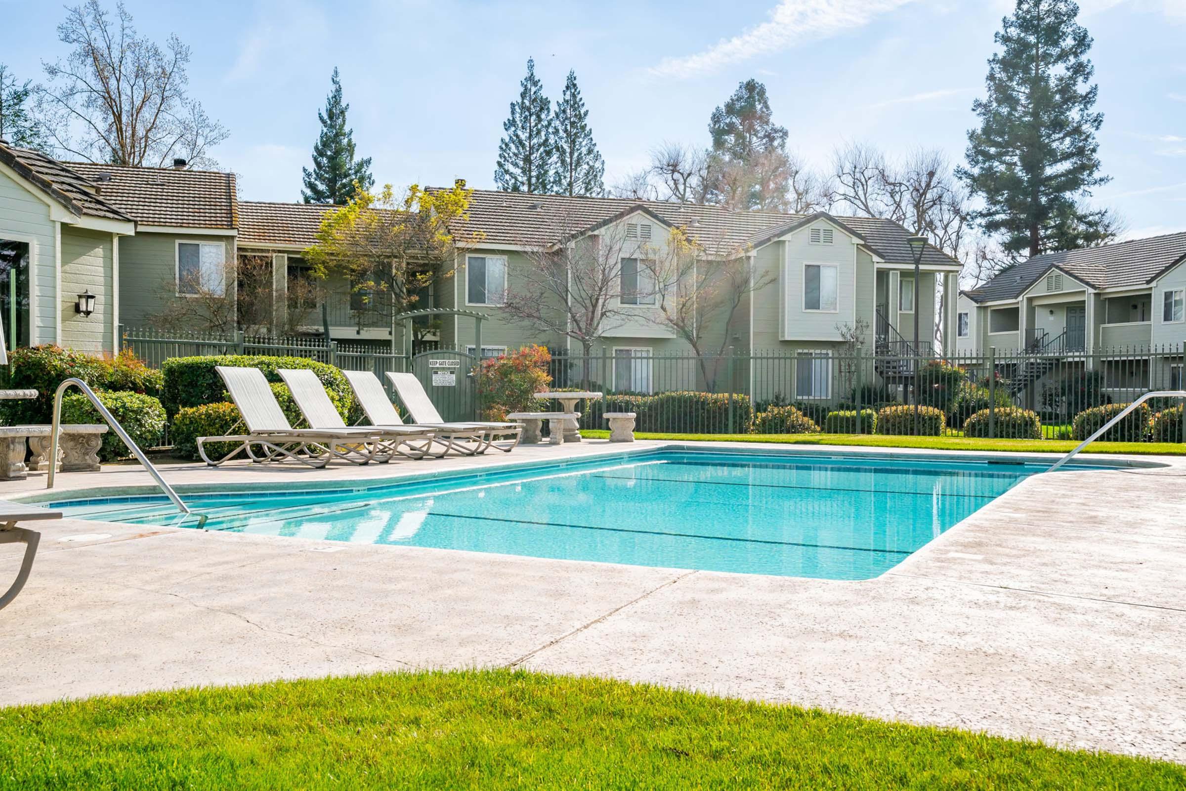 A clear swimming pool surrounded by lounge chairs, with well-maintained greenery and shrubs nearby. In the background, there are residential buildings with trees and a clear blue sky. The scene is sunny and inviting, perfect for relaxation.