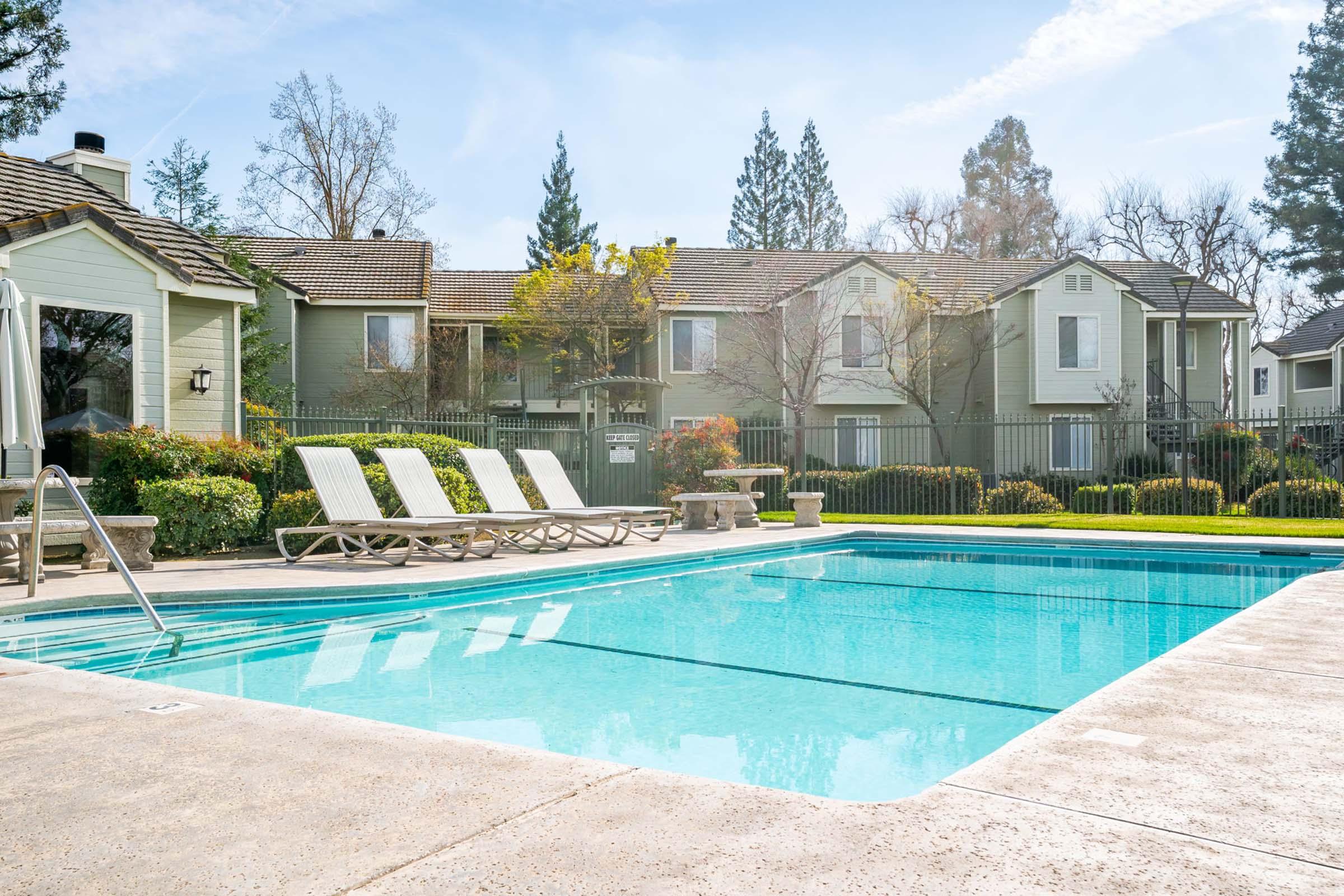 A serene swimming pool surrounded by lounge chairs, with a neatly landscaped area and trees in the background. The pool reflects a clear blue sky, while the adjacent buildings have a modern design and greenery nearby, creating a relaxing outdoor atmosphere.