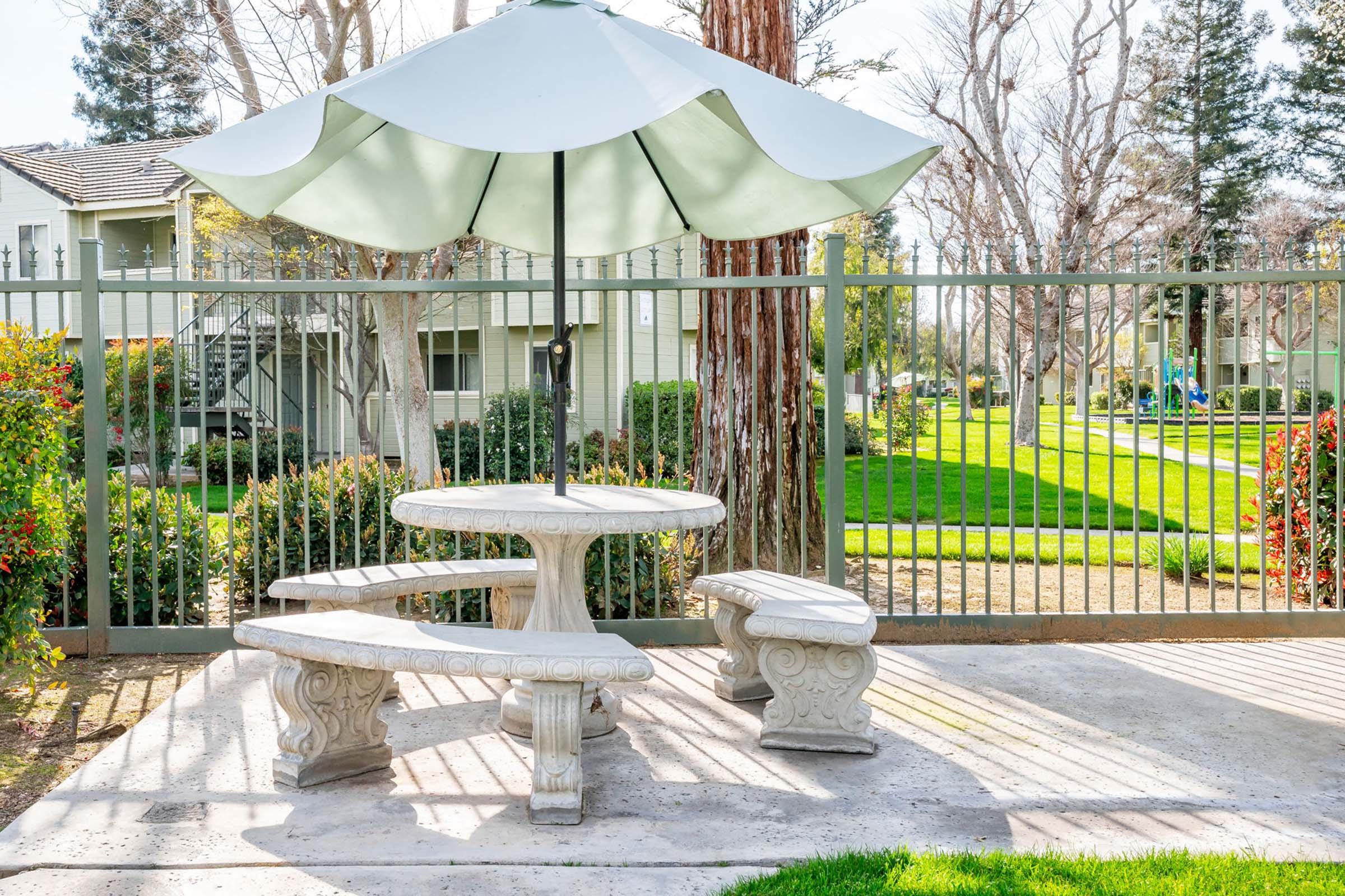 A stone picnic table with curved benches is shaded by a large, light-colored umbrella. The table is positioned in a grassy area surrounded by trees and a fence, offering a serene outdoor setting. Sunlight filters through the umbrella, creating gentle shadows on the ground.