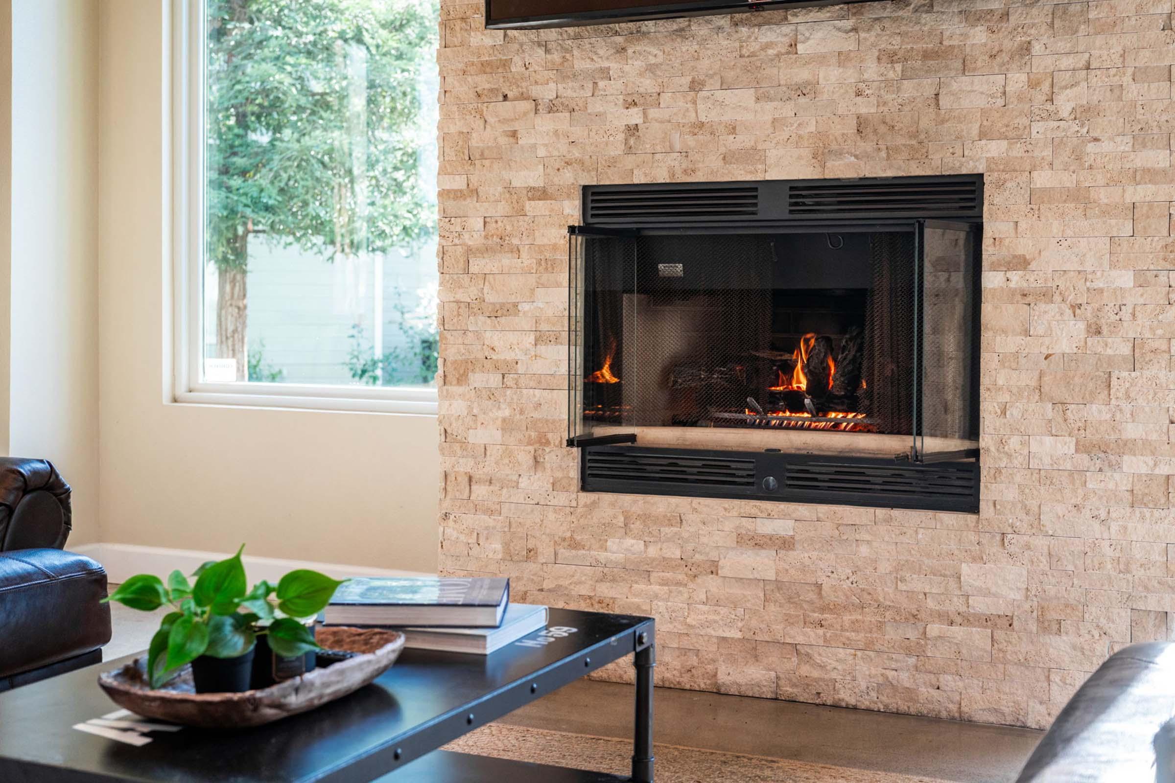 A cozy living room featuring a stone fireplace with a glass front, displaying a warm fire. A sleek black coffee table with a plant and a book sits nearby. Large windows allow natural light to fill the space, creating a welcoming atmosphere.