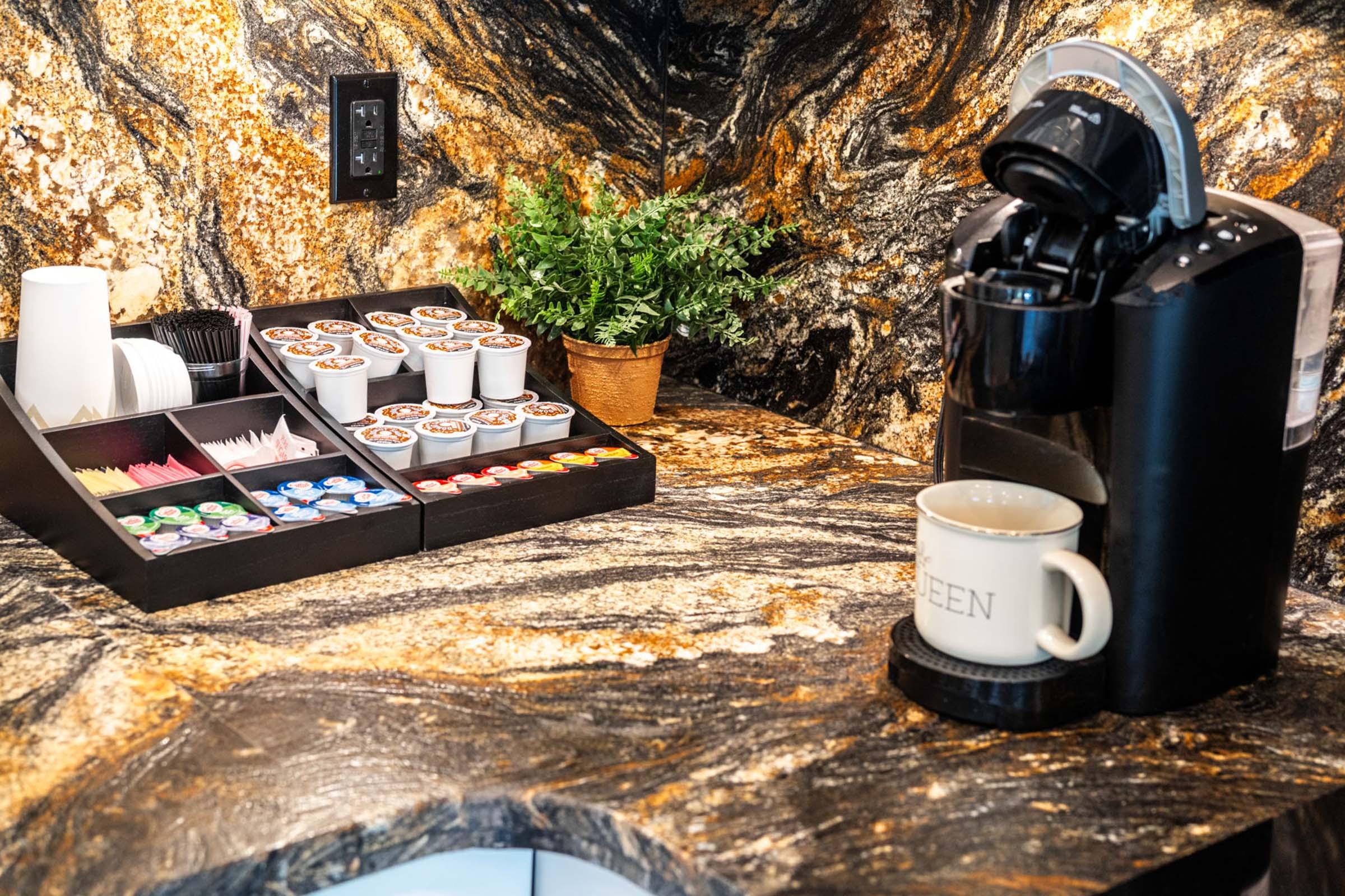 Coffee station featuring a coffee maker, a variety of coffee pods and creamers organized in a black tray, and a small potted plant on a textured granite countertop. A mug labeled "GREEN" is placed next to the coffee maker.