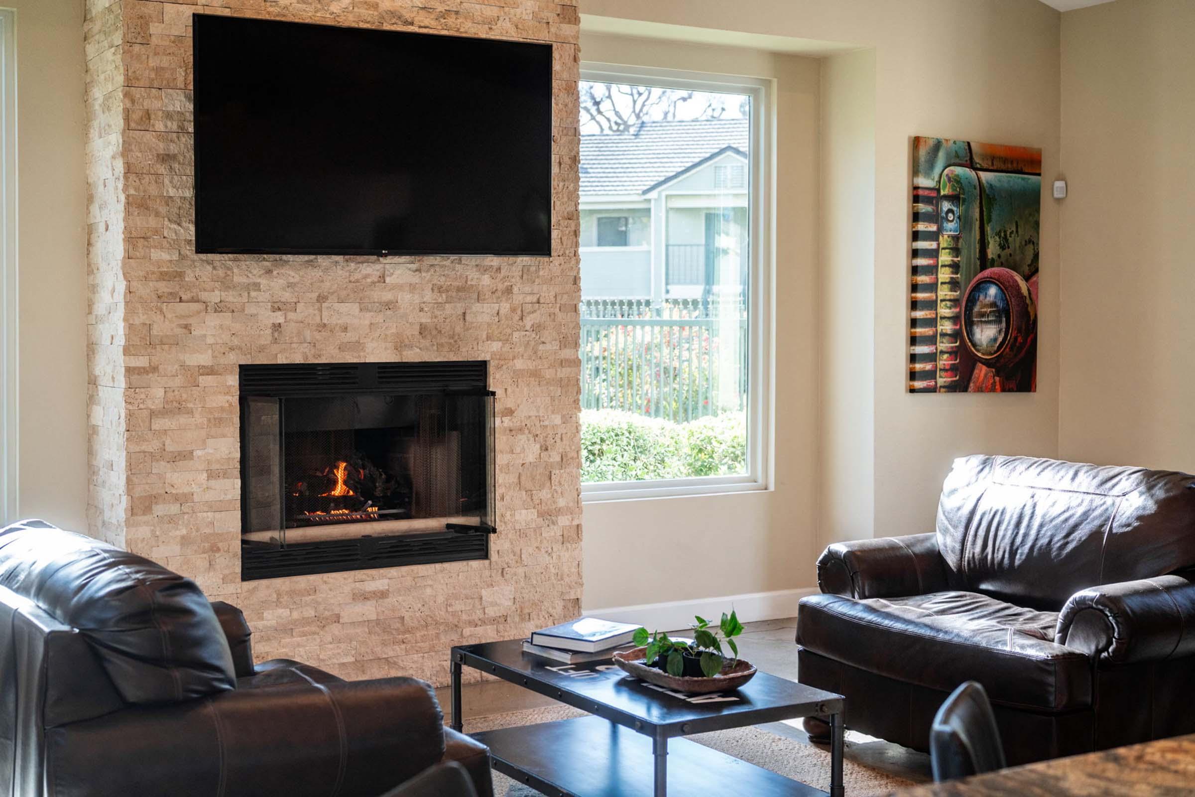 A cozy living room featuring a stone fireplace with a television mounted above it. Two brown leather armchairs are positioned in front of a coffee table with plants. A colorful painting of a vintage car adorns one wall, and large windows provide a view of the outdoors, adding natural light to the space.