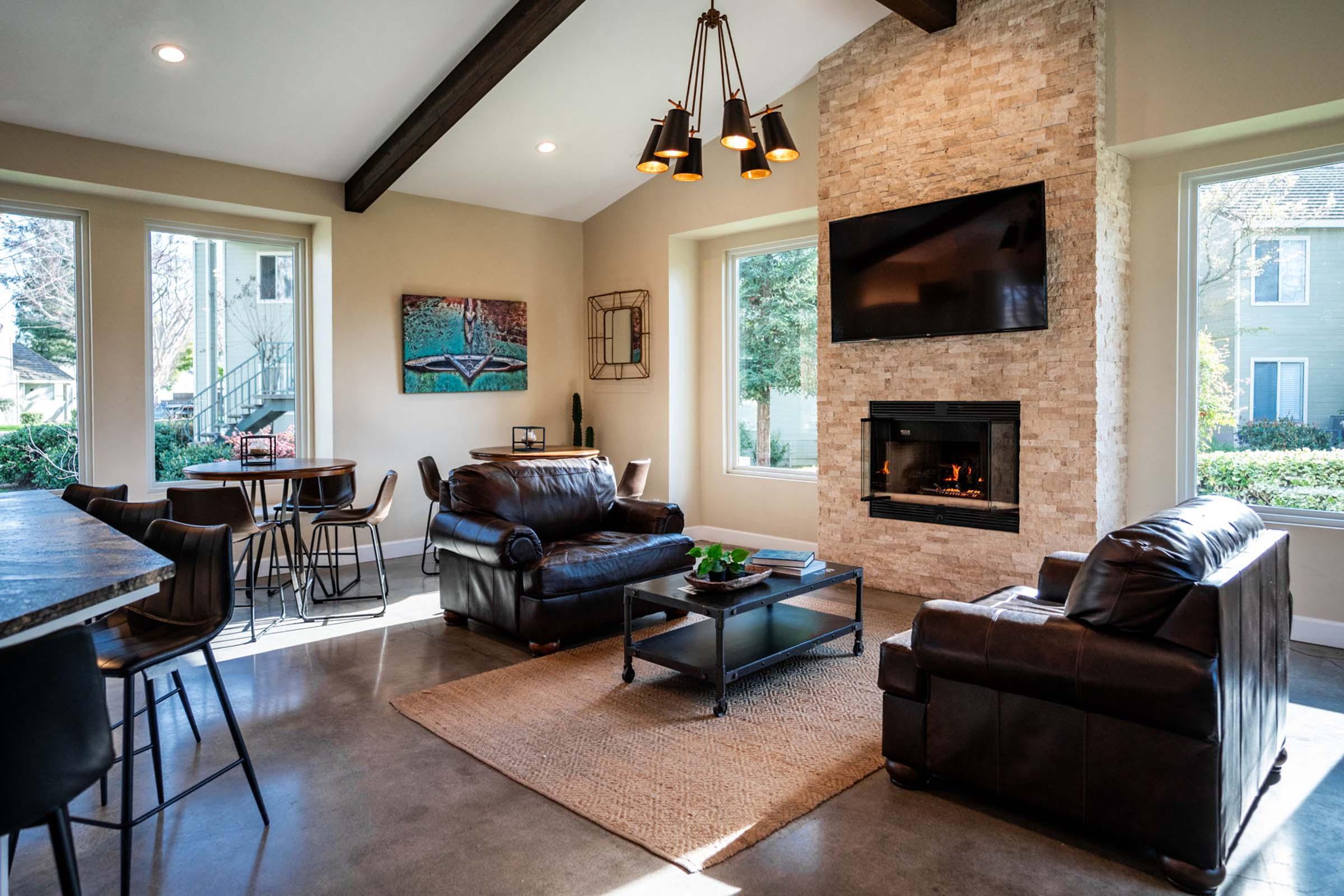 Cozy living room featuring a stone fireplace, a modern TV mounted on the wall, and large windows letting in natural light. There are two leather sofas, a coffee table on a textured rug, and a dining area with high chairs. The warm interior exudes comfort and style, with greenery visible outside the windows.