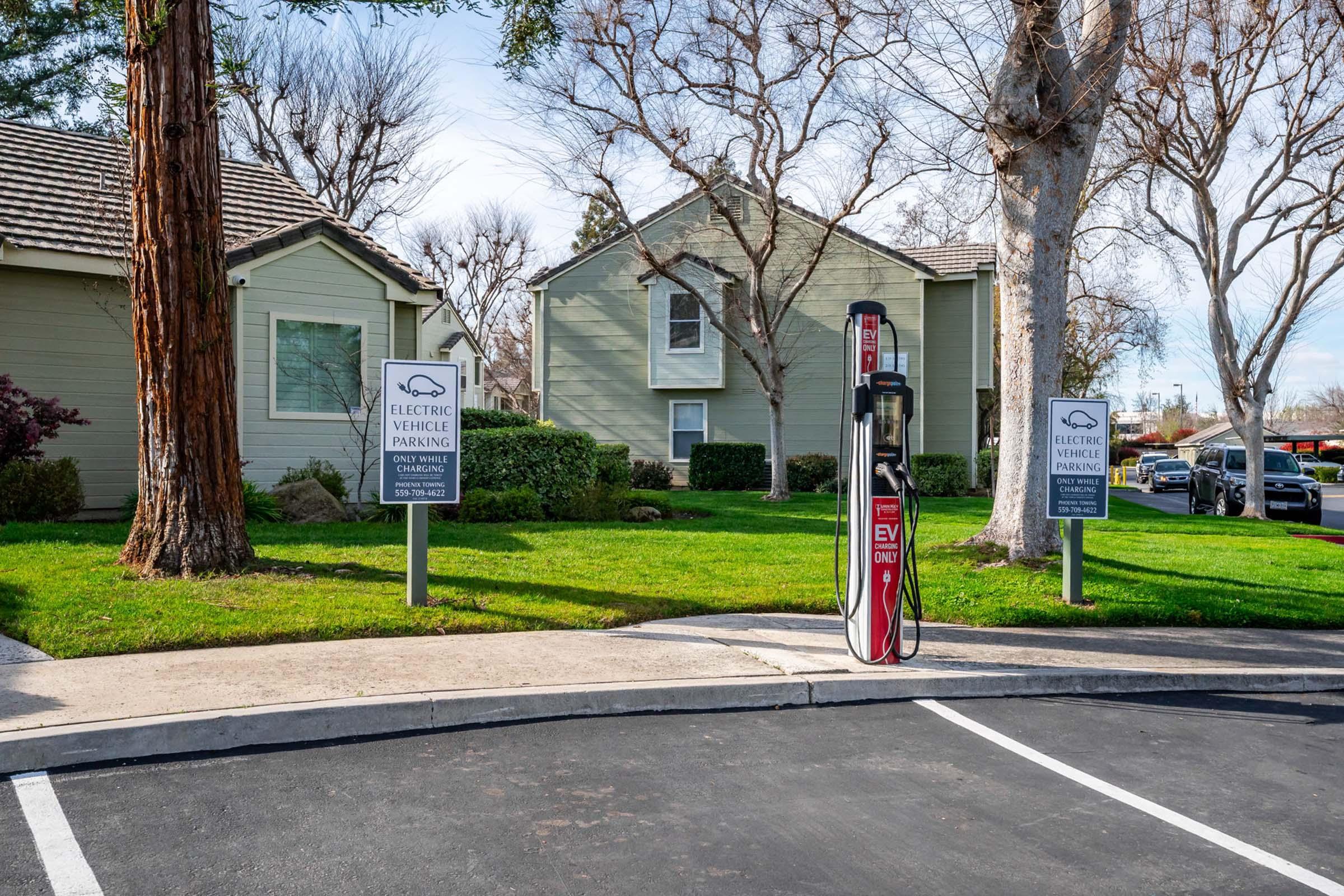 An electric vehicle charging station is located in a parking area next to two residential buildings. Signs indicate the space is reserved for electric vehicle parking only. There are trees and well-maintained grass surrounding the area, with additional vehicles parked nearby.